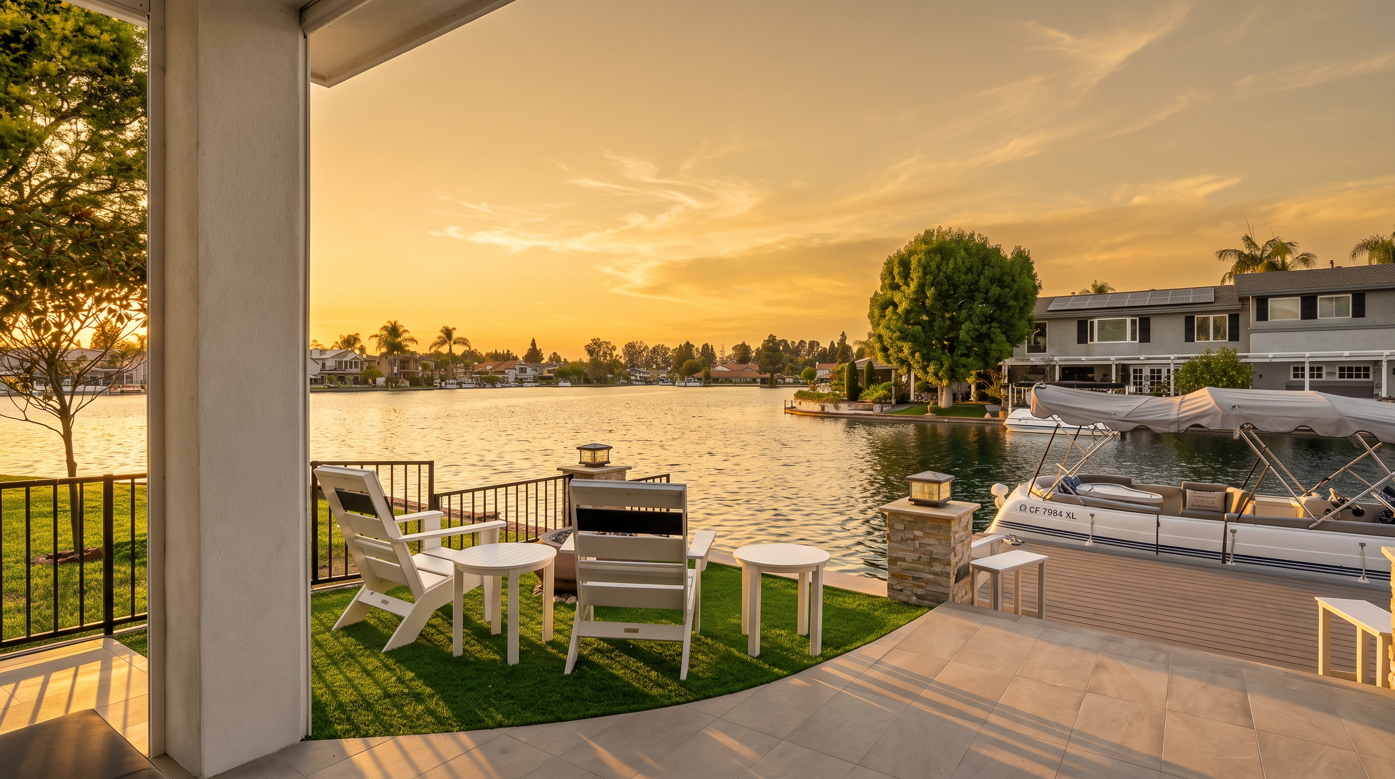 This stunning waterfront patio offers an inviting outdoor living space with a combination of tiled flooring and artificial turf. The area is furnished with stylish white chairs and small tables, perfect for relaxing while overlooking the serene lake and private boat dock. This setting creates an ideal, picturesque environment for leisure and entertaining in a tranquil lakeside community.