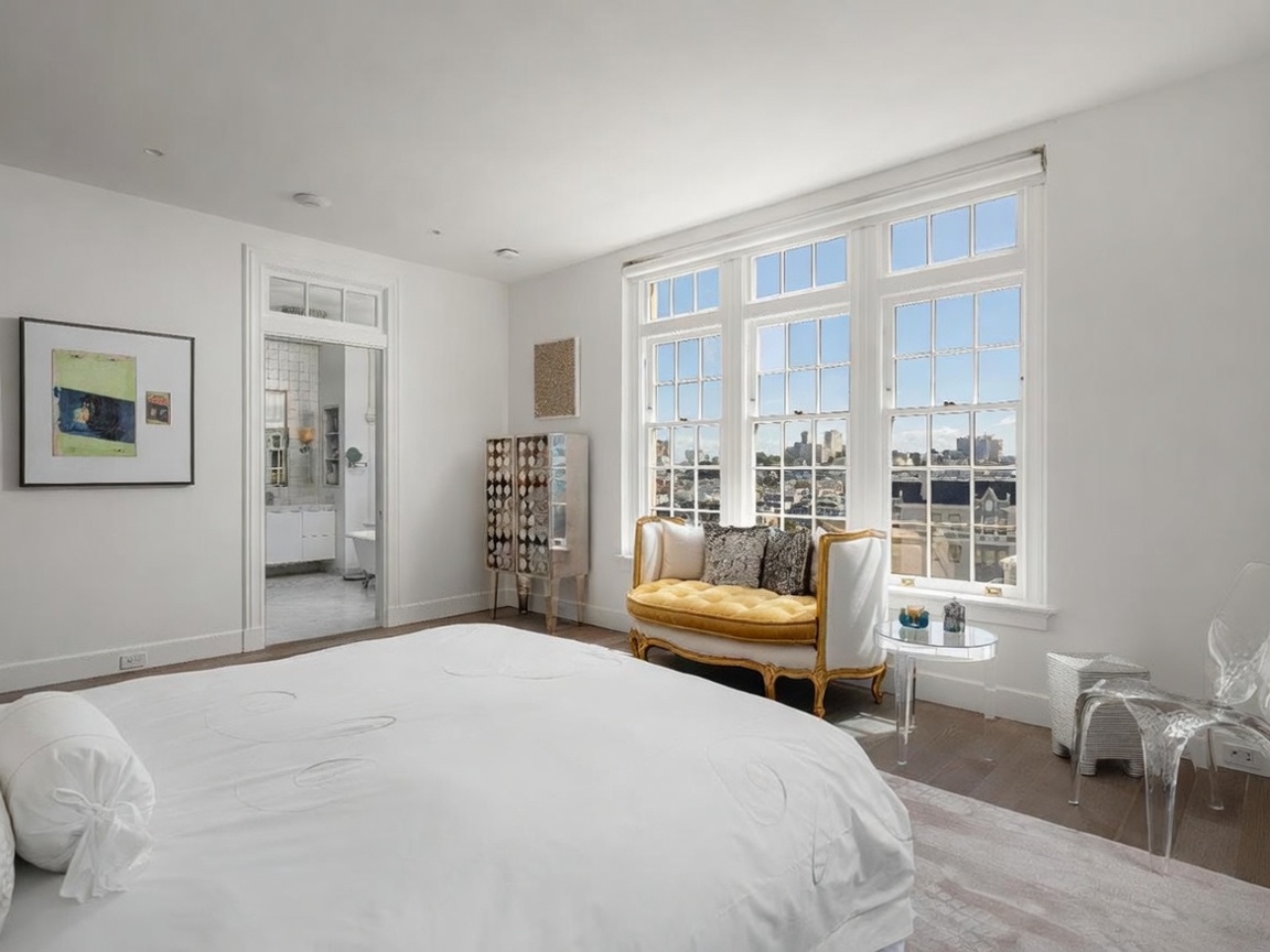 This bright and airy primary bedroom features a large, plush bed in the foreground and a sophisticated seating area positioned by a set of tall, multi-pane windows that offer an expansive city view. The room is decorated in a neutral palette with white walls, light wood flooring, and a decorative cabinet, while an open doorway leads into a bright, tiled bathroom. The perspective is captured from the foot of the bed, emphasizing the elegant transition between the sleeping area and the en-suite space.