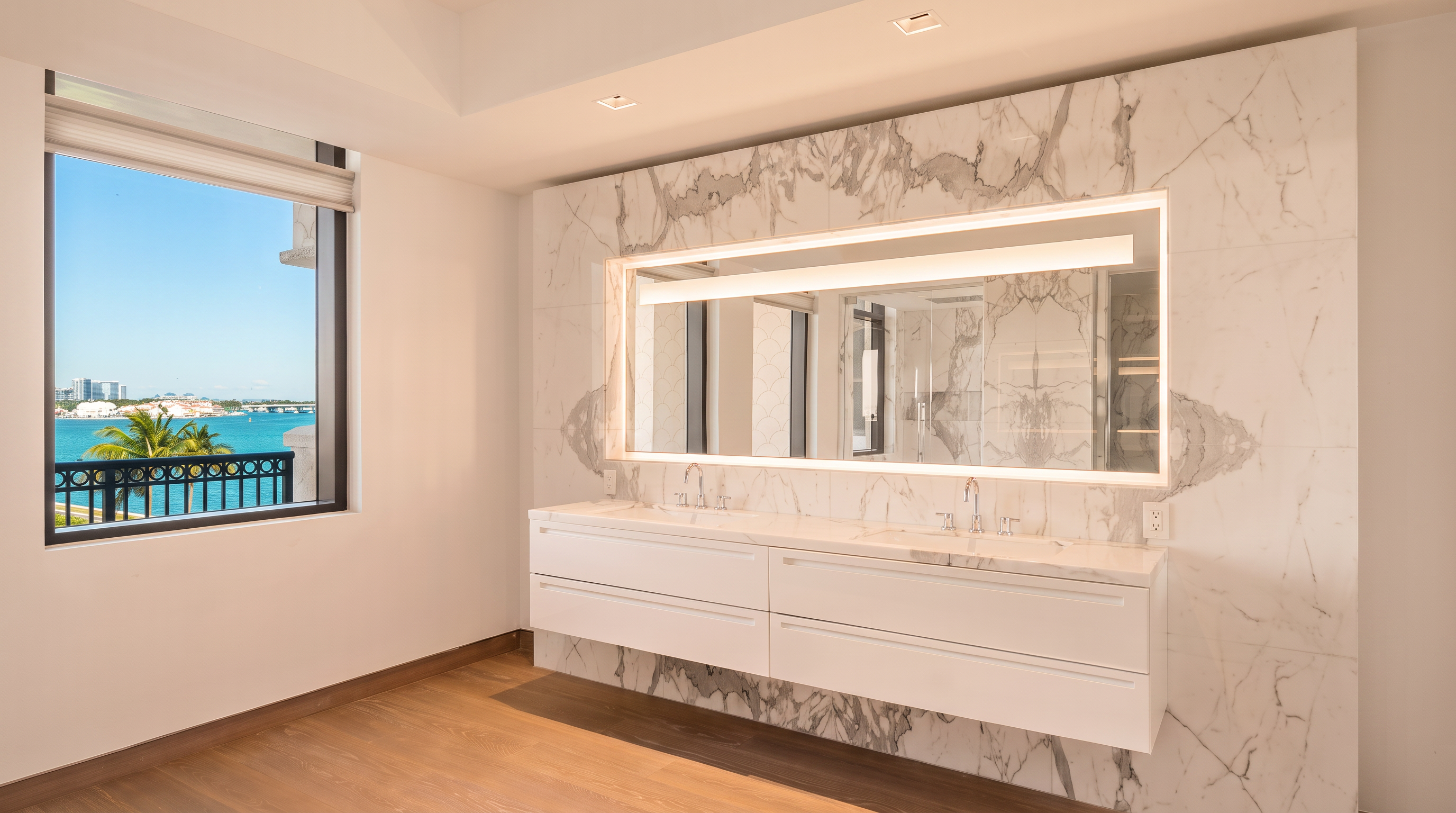 This modern primary bathroom features a sleek, wall-mounted double vanity with a clean white finish and integrated sinks. A large, rectangular mirror with integrated LED lighting is mounted against a striking, full-height stone wall with dramatic veining. To the left, a large window provides an expansive, unobstructed view of the ocean and sky, filling the space with natural light.