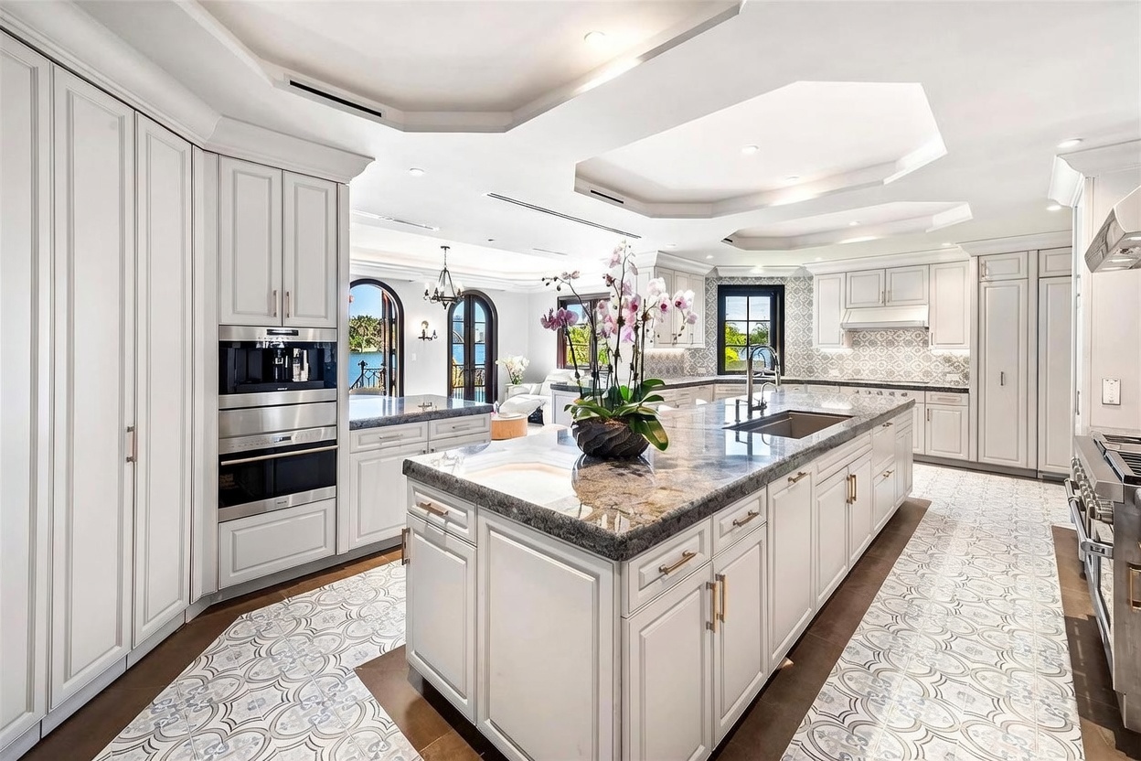 This expansive, high-end kitchen features white cabinetry, a large central island with a stone countertop, and intricate patterned tile flooring. The space is illuminated by recessed lighting and offers a clear view through to an adjacent living area with arched doorways overlooking the water. The design combines classic elegance with modern functionality, creating a bright and sophisticated atmosphere.