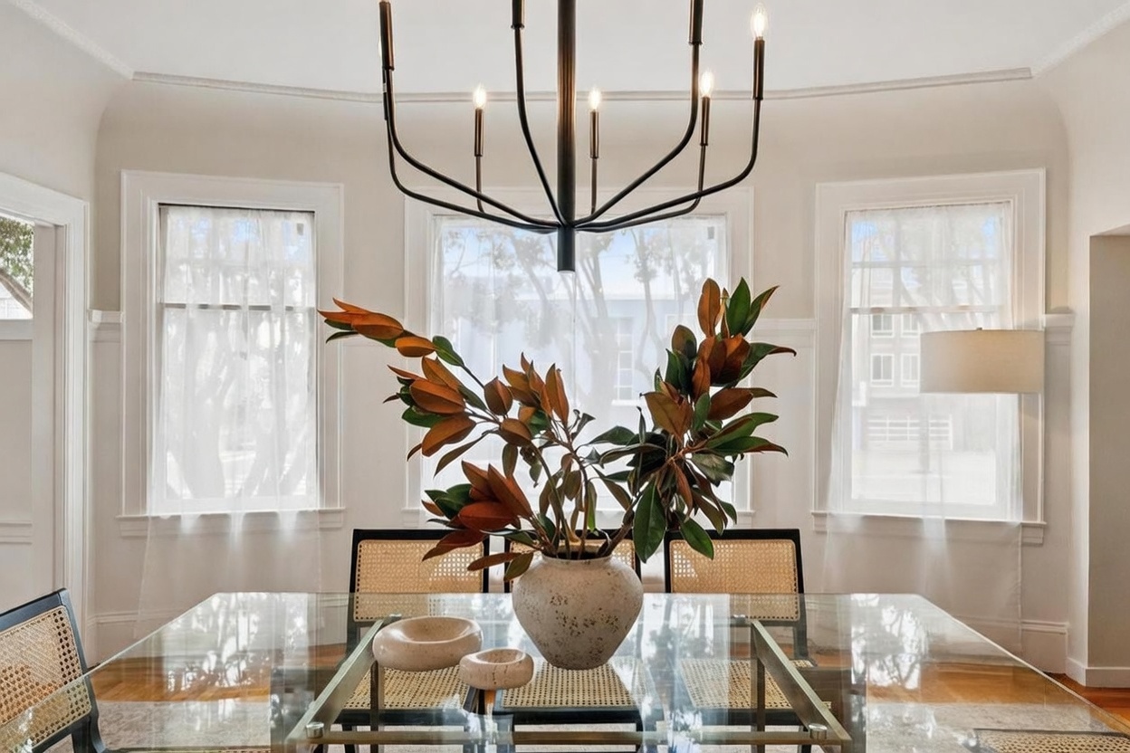 This elegant dining room features a glass-topped table surrounded by chairs with light-toned woven cane backs. A striking, modern black chandelier hangs above a central vase filled with large, glossy leaves, creating a sophisticated focal point. The room is brightened by three tall windows dressed in sheer white curtains, and a minimalist floor lamp stands in the corner, contributing to the clean, airy aesthetic.