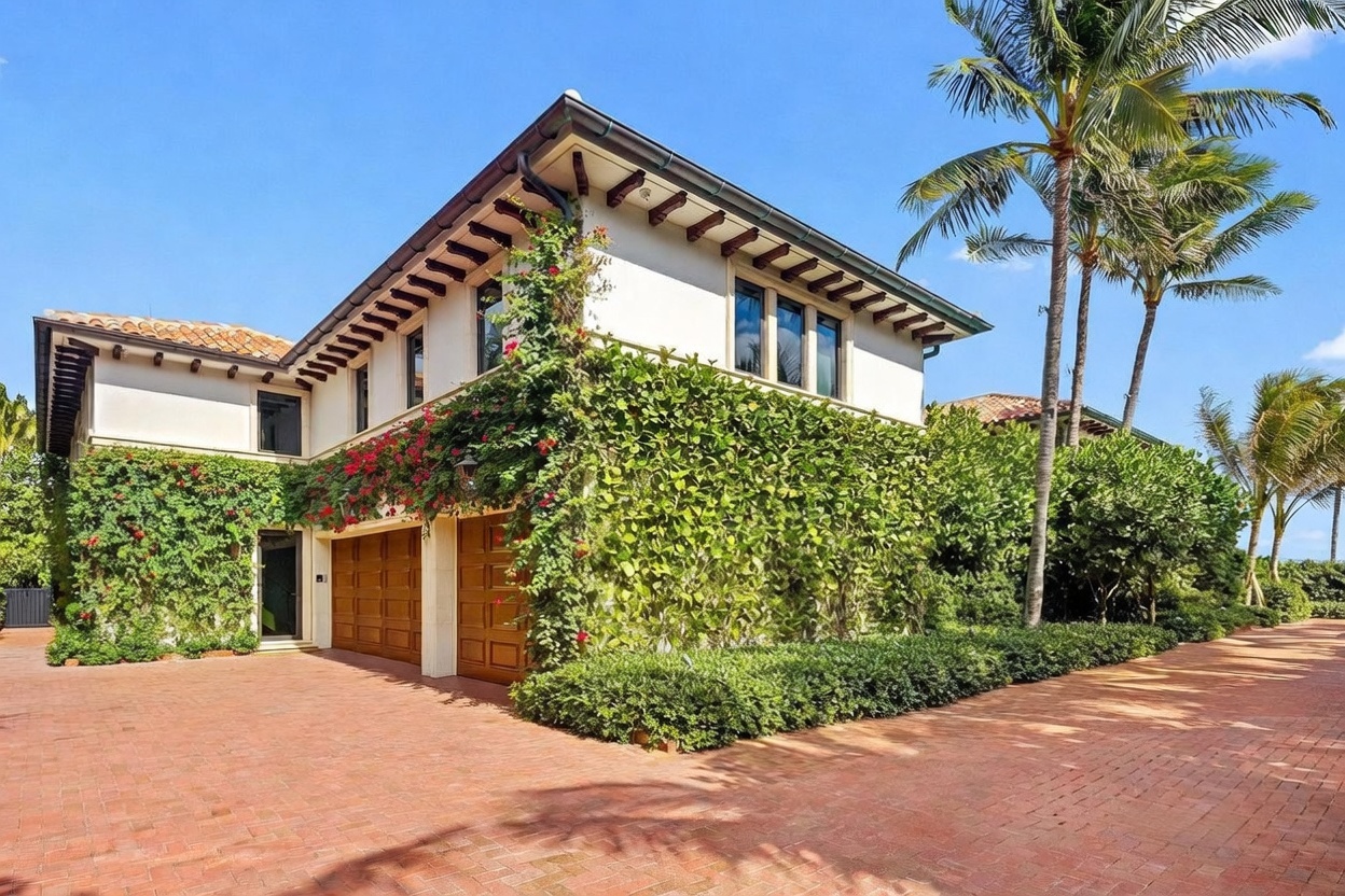 This Mediterranean-style home features a prominent garage with wooden doors, partially obscured by lush, flowering vines climbing the exterior walls. The property is approached by a wide, red brick-paved driveway, and tall palm trees frame the bright blue sky in the background. The architectural design includes decorative wooden corbels under the eaves and a warm-toned tile roof, creating a classic and inviting curb appeal.