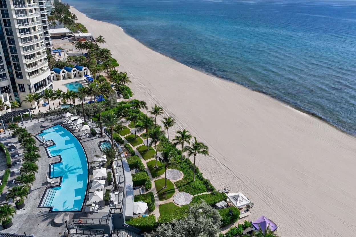 This high-angle aerial view captures a luxurious beachfront property featuring a sprawling, curved swimming pool and meticulously landscaped grounds. The scene showcases a seamless transition from the private resort-style amenities, including sun loungers and cabanas, to the expansive sandy beach and the vast blue ocean. The perspective highlights the prime coastal location and the sophisticated outdoor living spaces designed for relaxation and recreation.