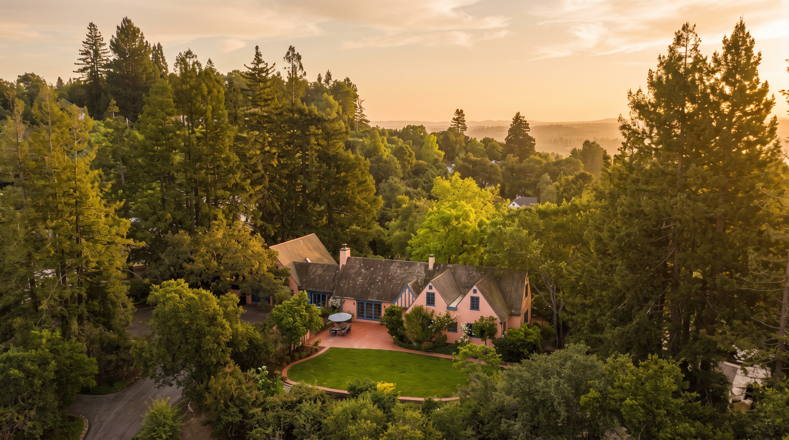 This high-angle aerial view captures a charming, multi-gabled home nestled within a lush, wooded landscape. The property features a terracotta-toned patio area adjacent to a manicured green lawn, surrounded by mature trees and rolling hills in the distance. The home's traditional architecture and warm exterior color palette create a serene and private atmosphere.