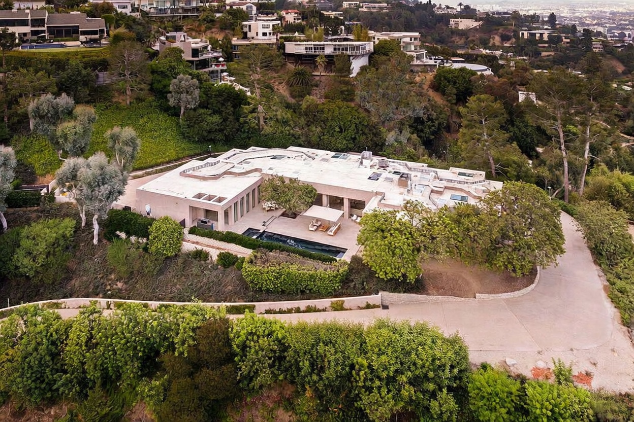 This high-angle aerial shot captures a sprawling, modern luxury estate nestled into a lush, hilly landscape. The property features a flat-roofed architectural design, a sleek rectangular swimming pool, and a spacious patio area surrounded by mature trees and manicured greenery. The perspective emphasizes the home's secluded, elevated position and its seamless integration with the surrounding natural terrain.