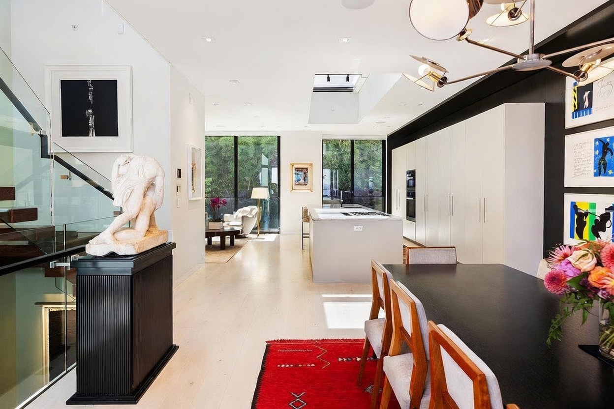 This open-concept interior space seamlessly blends a dining area with a modern kitchen and a distant living space. The foreground features a dark dining table paired with light-toned chairs, set against a wall adorned with framed artwork and a striking modern chandelier. A sculptural art piece sits on a black pedestal near a glass-railed staircase, while the kitchen beyond showcases a central island and large windows that provide a view of the lush greenery outside.