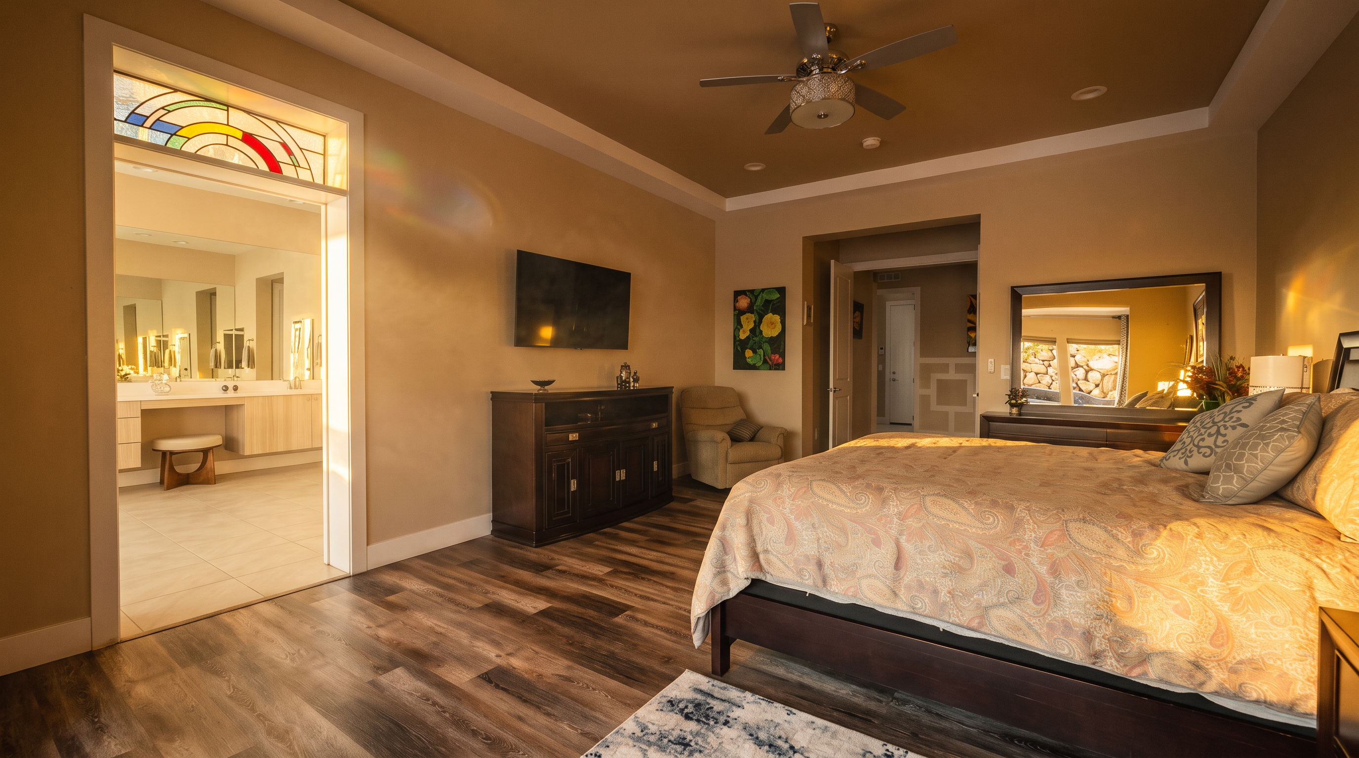 This spacious primary bedroom features wood-look luxury vinyl plank flooring and a tray ceiling with a central fan. The room is comfortably furnished with a bed, a dark wood dresser, a media console, and a cozy armchair, while providing a clear view into a well-appointed en-suite bathroom accented by a decorative stained-glass transom window.