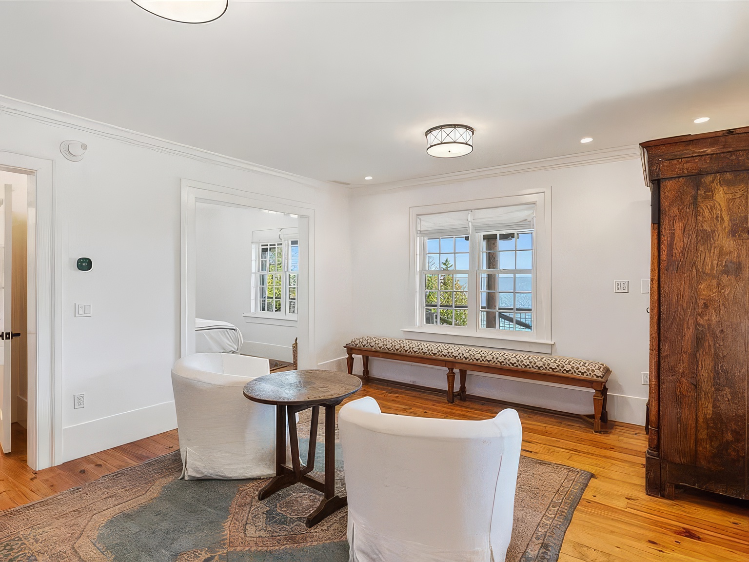This bright and airy sitting area features warm wood flooring, a patterned area rug, and two comfortable white armchairs centered around a small round wooden table. A long, upholstered bench sits beneath a large window that offers a scenic view of the water, while a tall, dark wood antique armoire adds a touch of traditional character to the space. The room connects to an adjacent bedroom through a wide doorway, creating an open and inviting atmosphere.