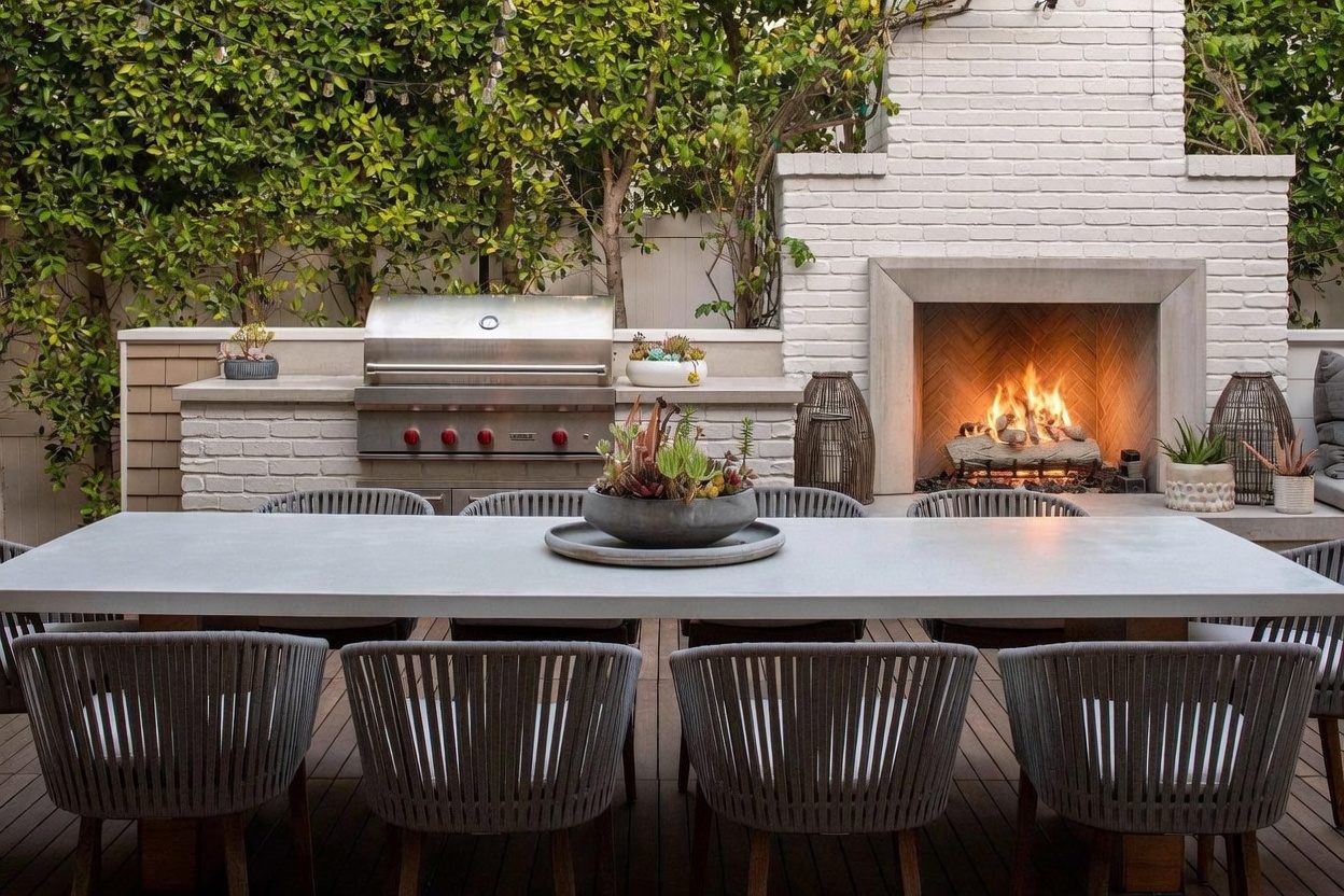 This inviting outdoor dining area features a long, sleek table surrounded by modern, woven-back chairs, set against a backdrop of a lush green hedge. A built-in stainless steel grill is integrated into a brick-faced outdoor kitchen counter, positioned next to a tall, white-painted brick fireplace with a glowing fire. The scene is captured from a low, eye-level perspective, emphasizing the cozy and sophisticated atmosphere of this private patio space.