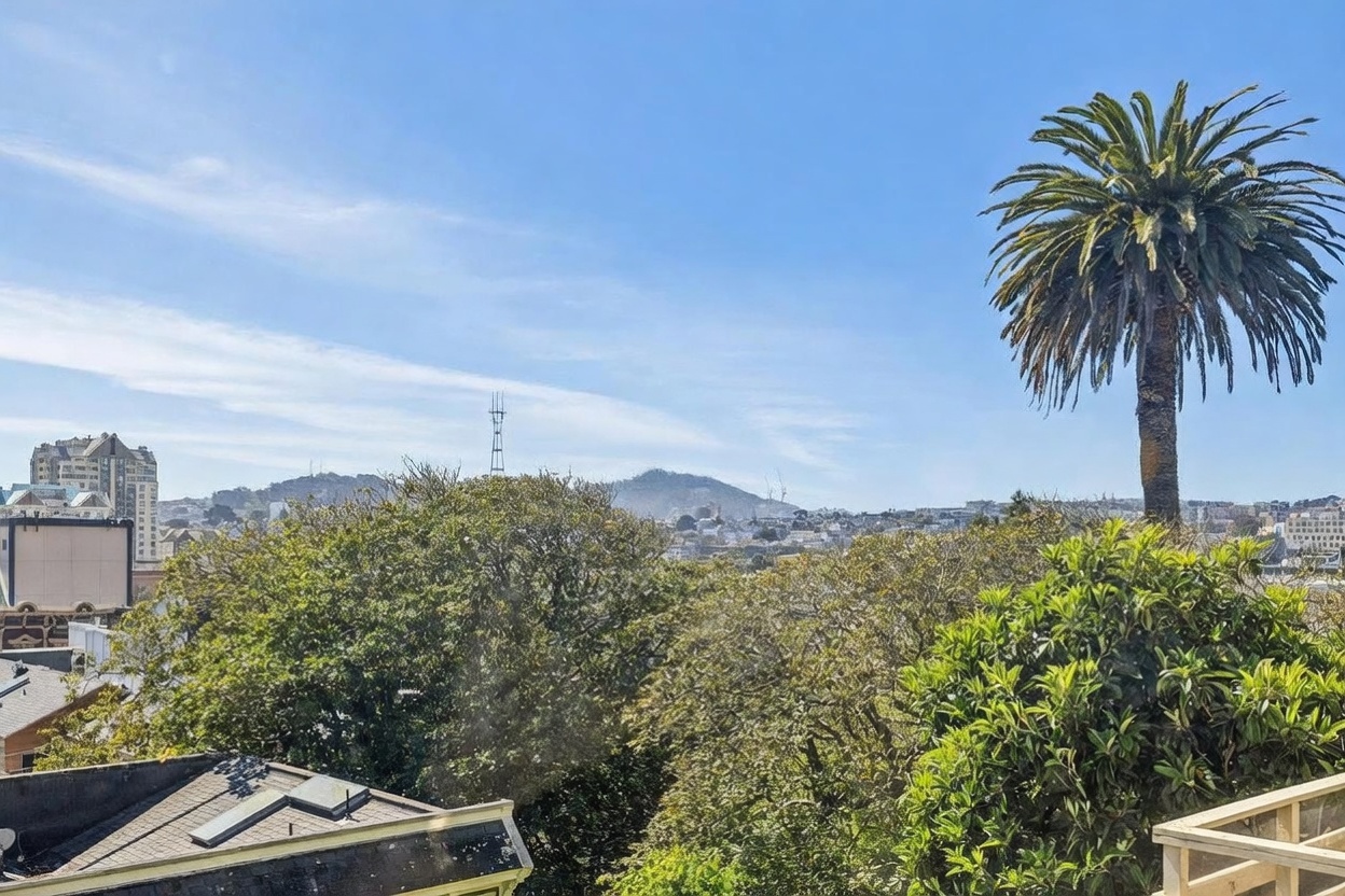 This view from an elevated balcony captures a scenic cityscape under a clear blue sky. The foreground features lush green trees and a prominent palm tree, while the background showcases rolling hills and urban architecture, including a distant radio tower. The perspective provides a sense of height and openness, highlighting the property's desirable vantage point.