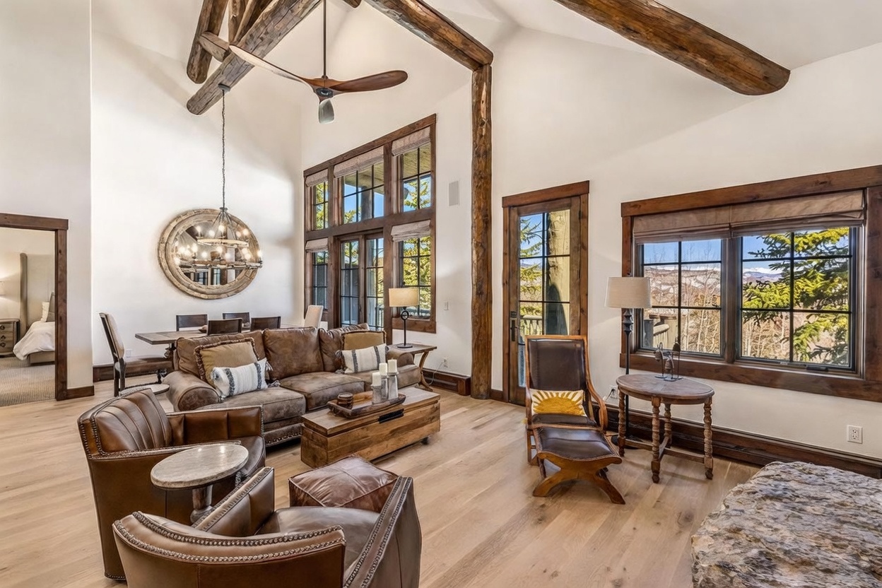 This expansive living room features a rustic mountain aesthetic with vaulted ceilings, exposed timber beams, and light wood flooring. The space is anchored by a large brown leather sofa and matching armchairs, complemented by a wooden coffee table and a view of snow-capped mountains through large windows. A dining area with a chandelier is visible in the background, creating an open-concept layout perfect for entertaining.