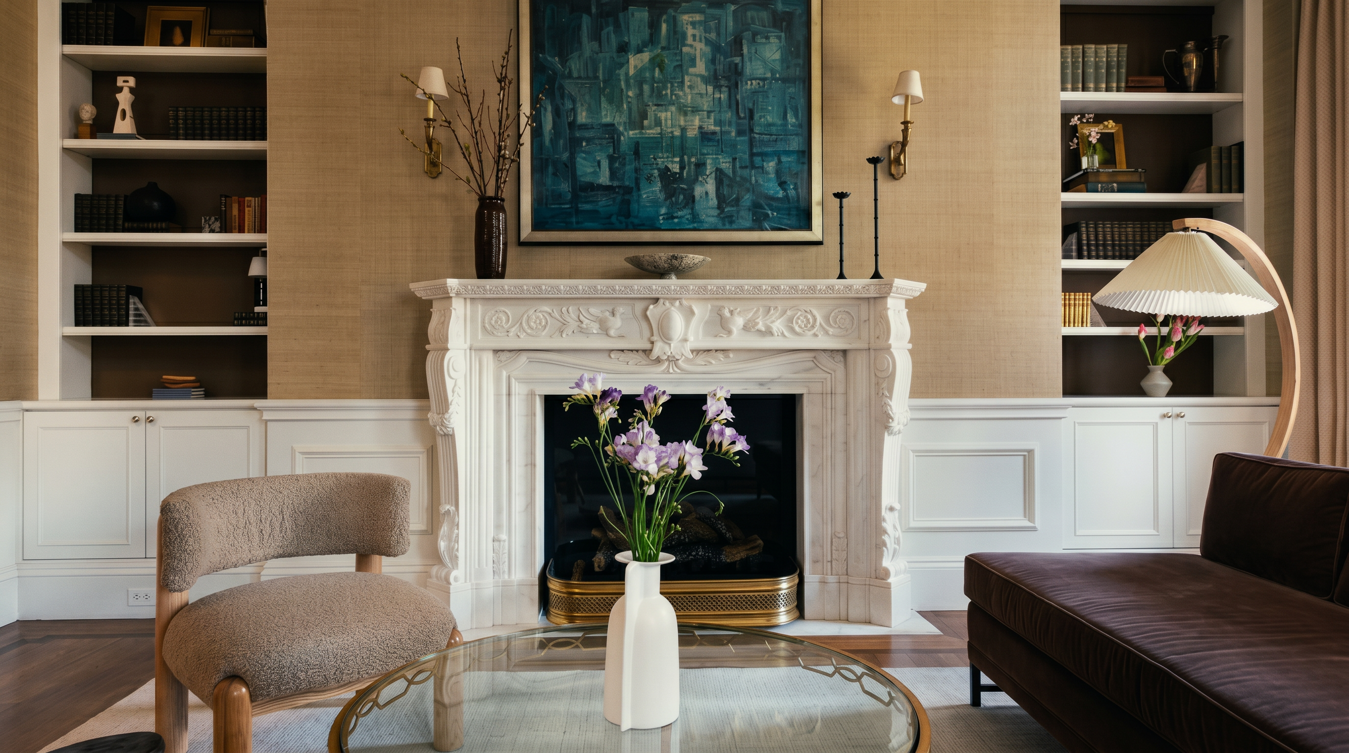 This elegant living room features a prominent stone fireplace surround as the central focal point, topped with a large abstract painting. Built-in shelving units flank the fireplace, providing ample display space, while a textured, neutral-toned wall covering adds warmth to the room. A glass-topped coffee table sits in the foreground, complemented by a textured armchair and a plush brown sofa, creating a sophisticated and inviting atmosphere.