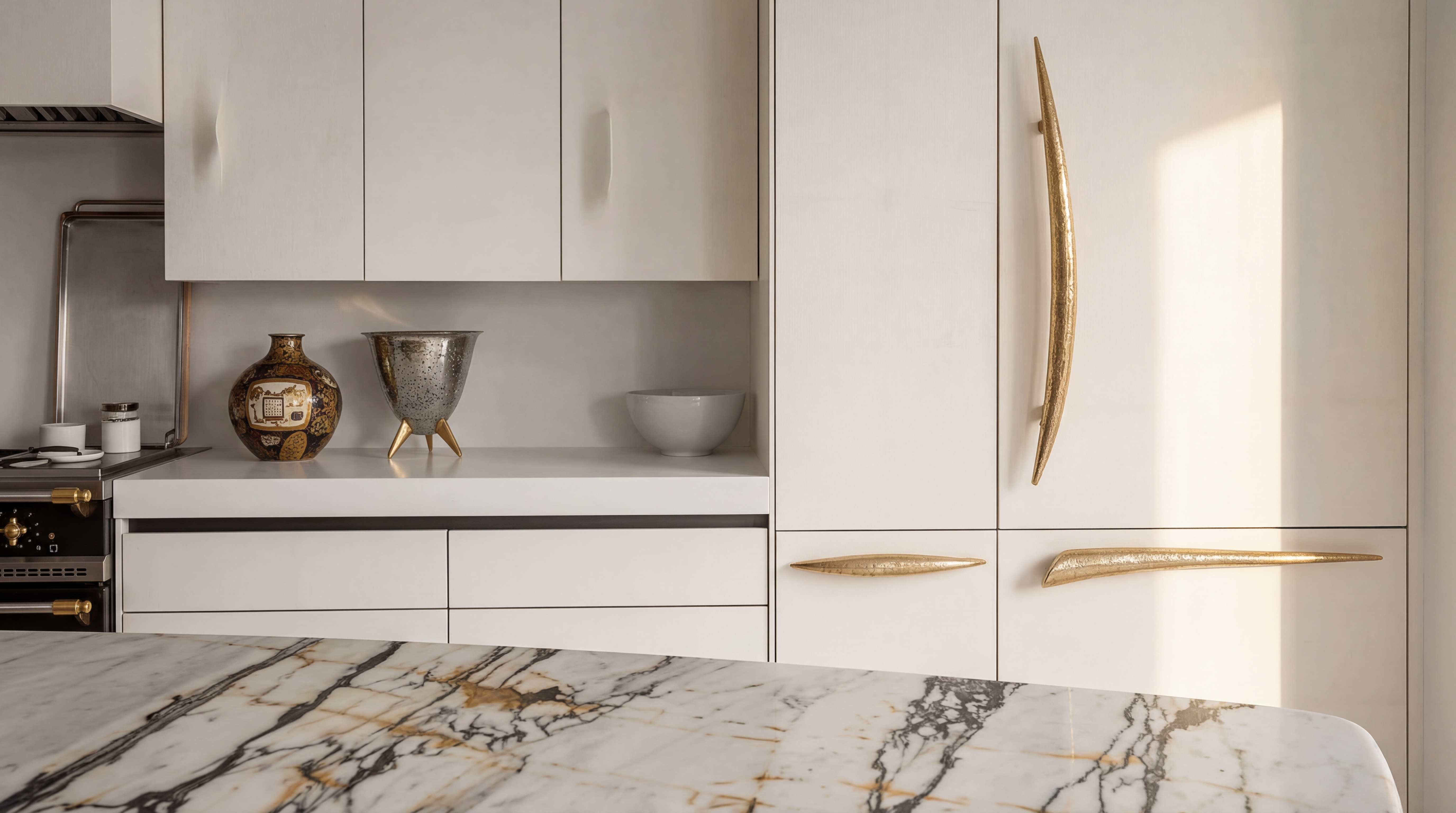 This sophisticated kitchen features clean, modern white cabinetry accented by striking, organic-shaped gold hardware. The focal point is a luxurious marble countertop with dramatic black and gold veining that complements the elegant decorative objects on the shelf. The space offers a high-end, contemporary aesthetic, highlighted by warm, natural light that enhances its premium design.