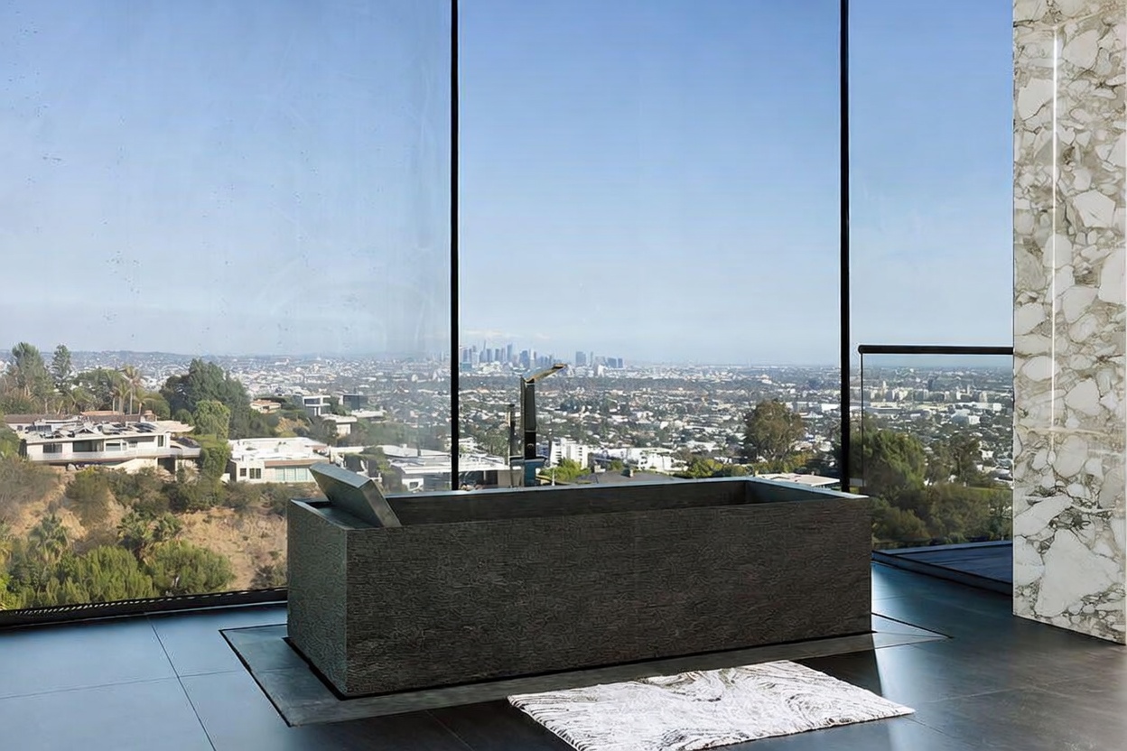 This luxurious primary bathroom features a striking, dark textured stone soaking tub positioned as the centerpiece against a floor-to-ceiling glass wall. The expansive window offers a breathtaking, panoramic view of the city skyline and surrounding hills, creating a serene and sophisticated atmosphere. A marble-clad pillar and a patterned rug on the dark tiled floor add elegant textures to this modern, high-end space.