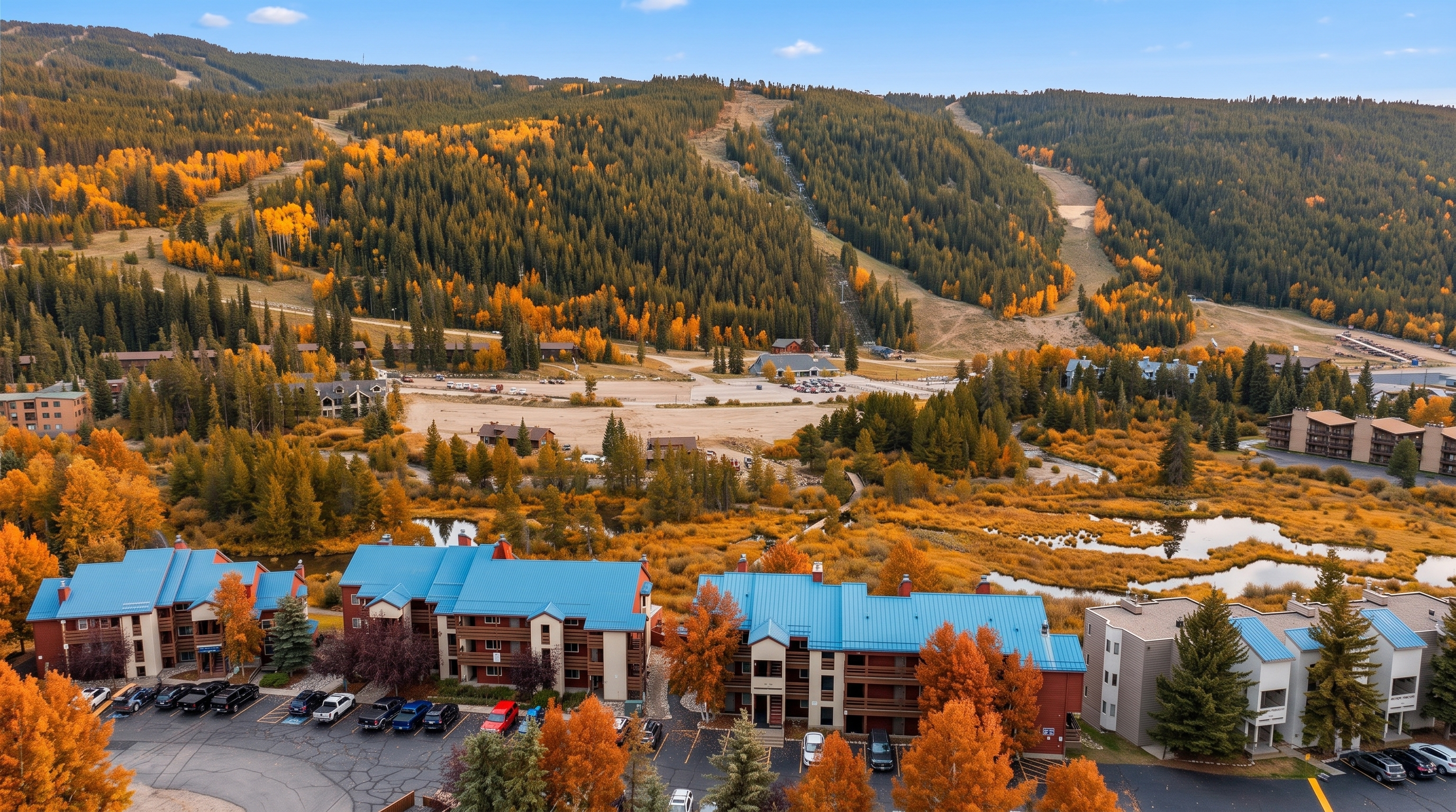 This stunning aerial view captures a scenic resort complex nestled in a vibrant, autumn-foliaged mountain landscape. The property features buildings with distinctive blue roofs, ample parking, and peaceful marshy ponds, all set against a backdrop of dense, golden-hued evergreen forests and mountain slopes. The image showcases the property's prime location, offering a tranquil and picturesque setting perfect for seasonal recreation.