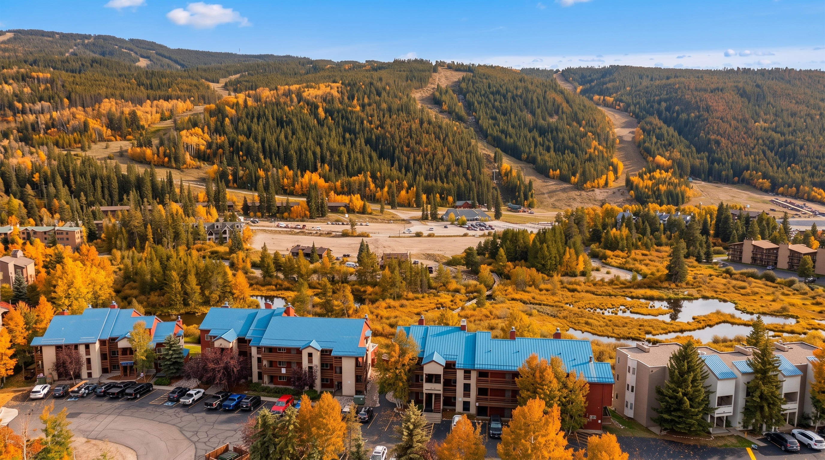 This aerial view captures a scenic multi-family mountain resort complex nestled in a vibrant autumn landscape. The buildings feature distinctive blue metal roofs and are surrounded by colorful fall foliage, a winding stream, and a backdrop of expansive evergreen mountains. The setting offers a peaceful and picturesque retreat environment, highlighting the property's integration with the natural landscape.