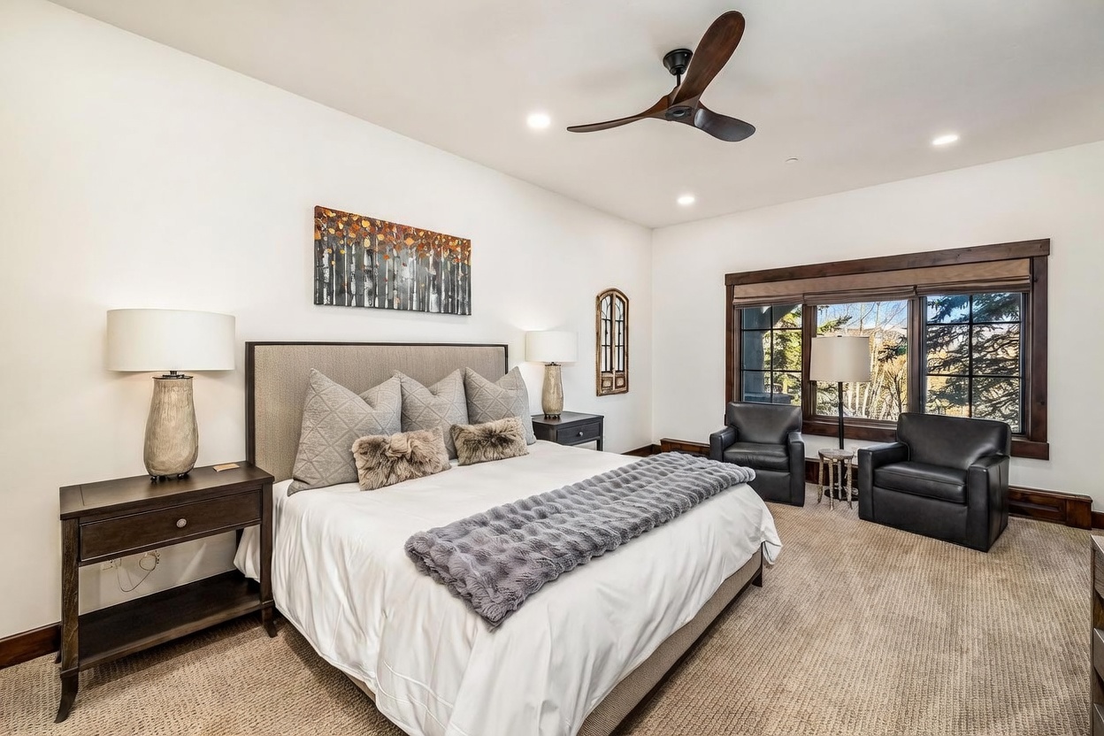 This spacious primary bedroom features a large bed with a neutral upholstered headboard, accented by textured pillows and a plush gray throw blanket. The room is furnished with dark wood nightstands, matching table lamps, and a pair of leather armchairs positioned near large windows that offer a view of the outdoors. A ceiling fan hangs above, and a piece of forest-themed wall art adds a decorative touch to the bright, clean space.