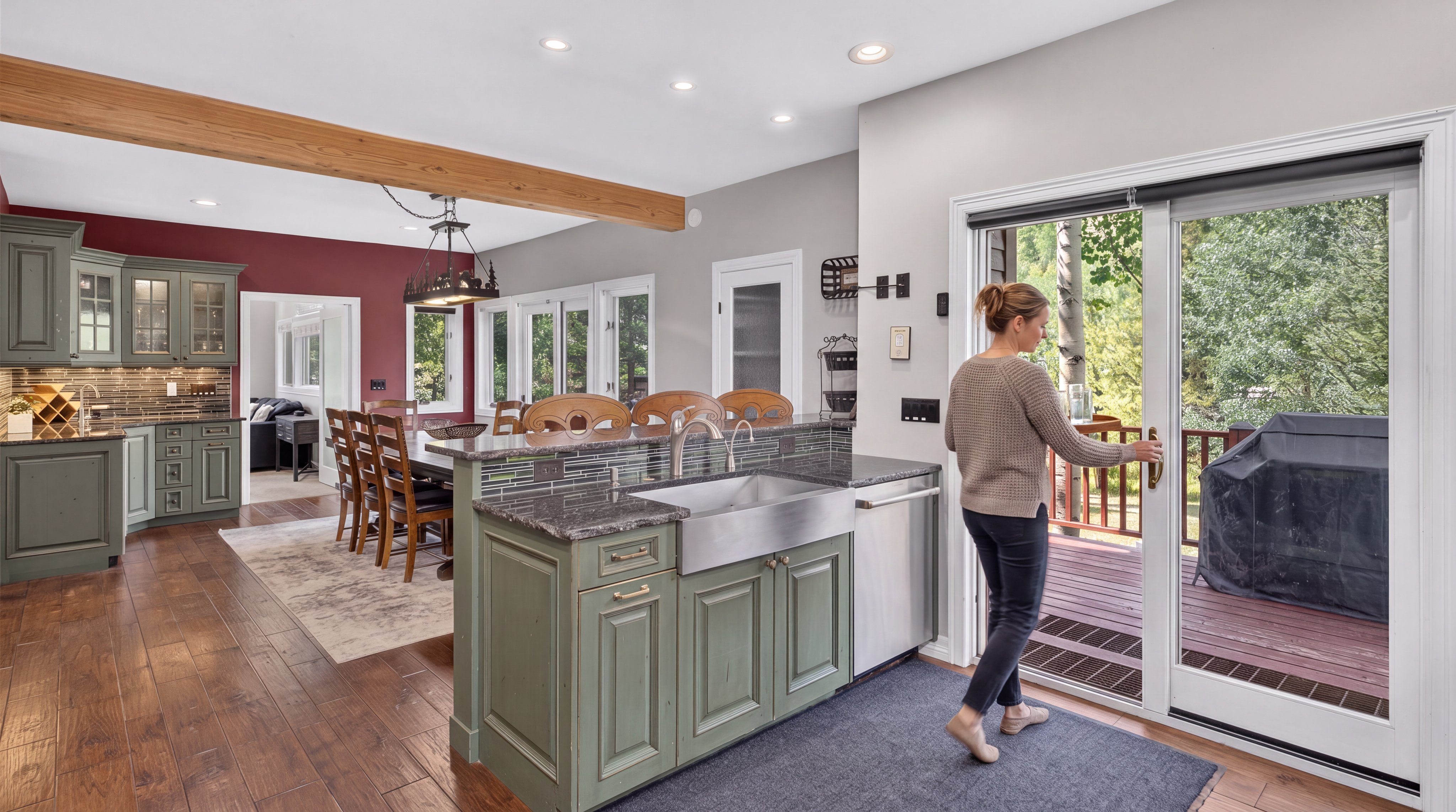 This kitchen features a striking blend of rustic and modern elements, including sage green cabinetry, a deep farmhouse sink, and elegant dark granite countertops. A wood-beamed ceiling and hardwood floors add warmth, while an open floor plan connects the kitchen seamlessly to a formal dining area and an outdoor deck through a large sliding glass door. The space is bright and inviting, perfect for entertaining and daily living.