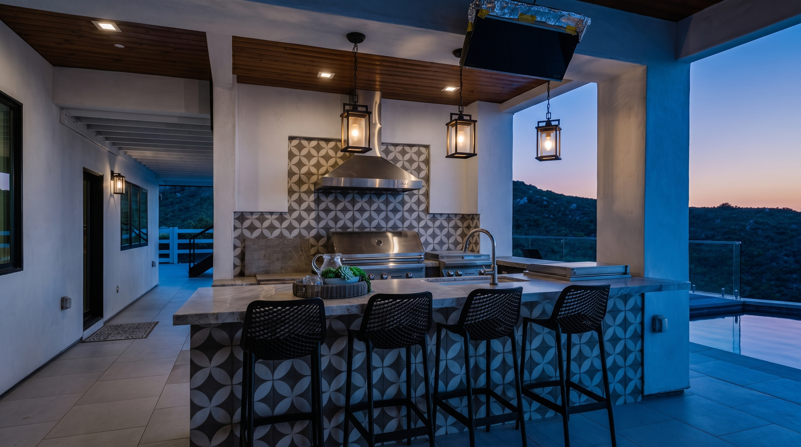 This luxurious outdoor kitchen and bar area is perfect for alfresco dining, featuring high-end stainless steel appliances, a modern patterned tile facade, and chic black bar stools. The space is covered by a wooden ceiling with hanging lanterns and opens up to an expansive terrace overlooking a pool and stunning scenic hills. It offers a seamless blend of contemporary design and functional outdoor living.