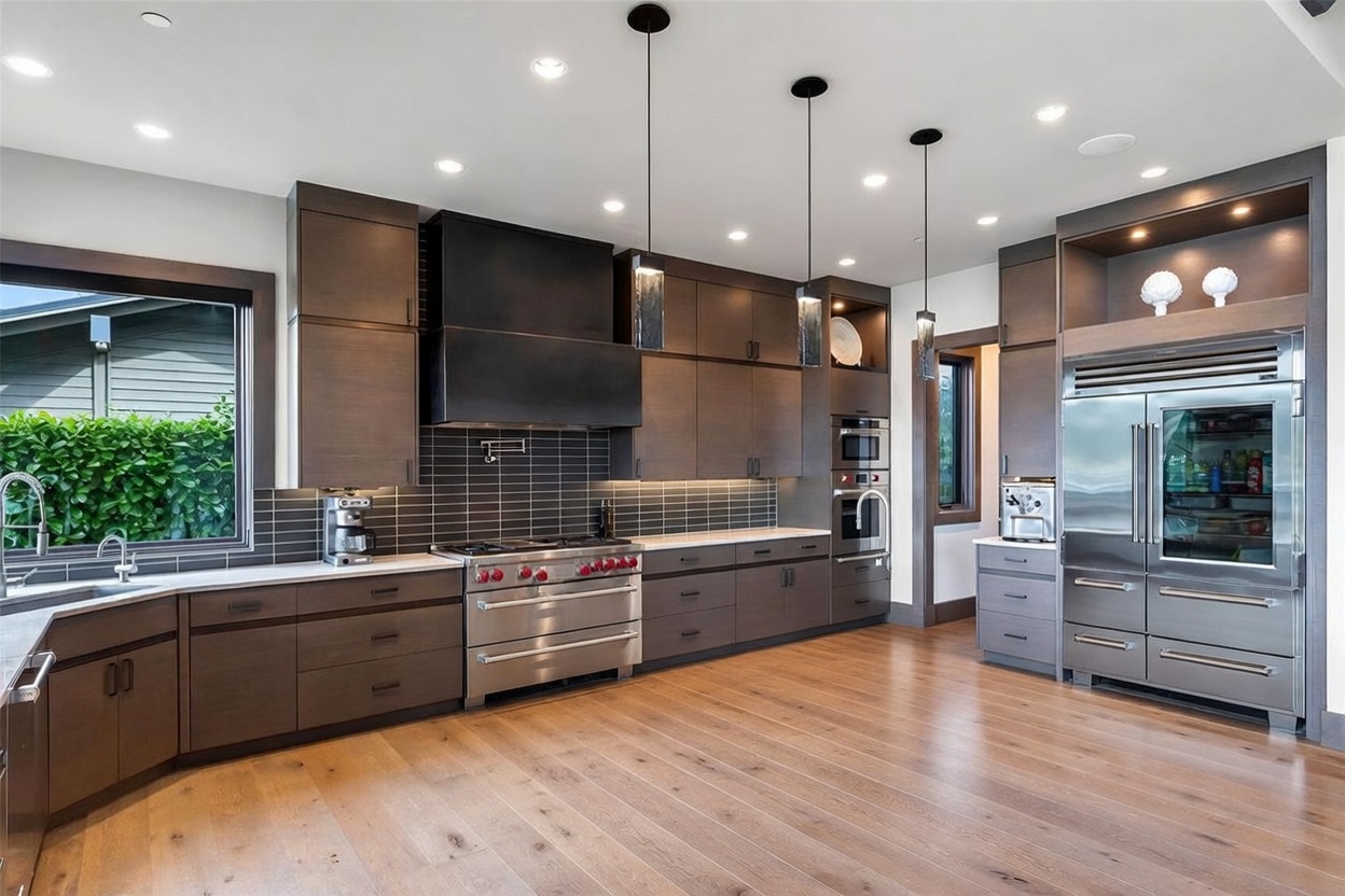 This modern kitchen features dark wood cabinetry, a large central island with a stone countertop, and a professional-grade range with a matching dark hood. The space is illuminated by three sleek pendant lights hanging over the island and recessed lighting throughout the ceiling. A tiled backsplash adds texture to the wall, while large windows provide natural light and a view of the outdoors.