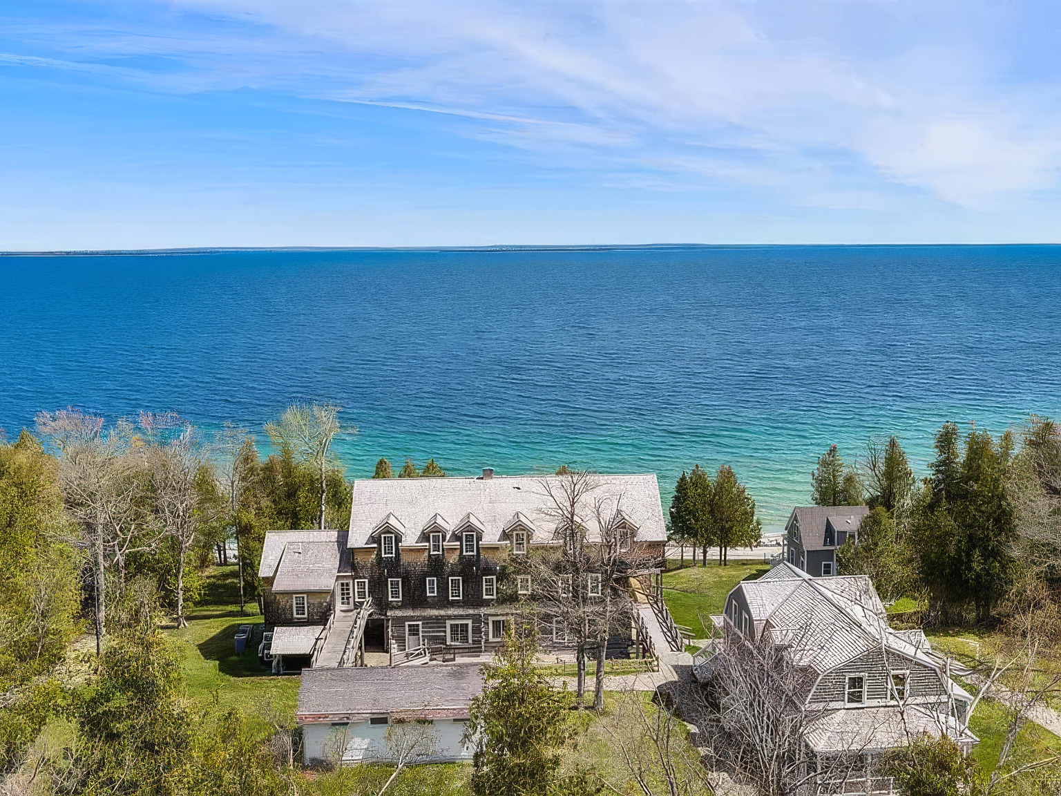 This high-angle aerial view captures a large, rustic-style multi-story residence situated on a lush, tree-lined property overlooking a vast, deep blue body of water. The main structure features a weathered shingle exterior with multiple dormer windows and a prominent roofline, complemented by a smaller, separate building in the foreground and a secondary house to the right. The expansive waterfront setting and mature trees create a serene, secluded atmosphere ideal for a coastal or lakeside retreat.