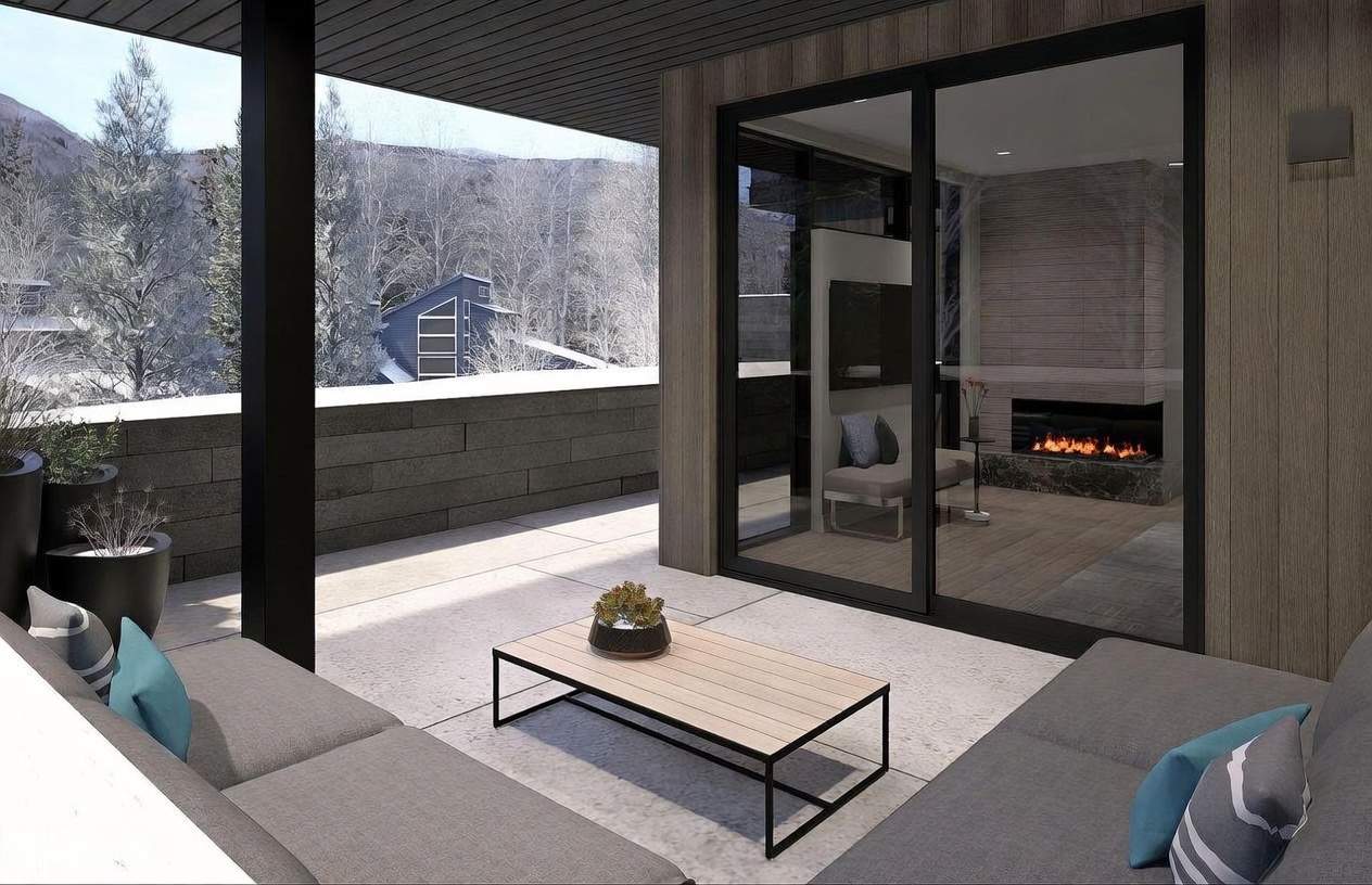 This modern balcony offers a seamless transition between indoor and outdoor living, featuring a comfortable seating area with plush gray sofas and a central coffee table. Through the large sliding glass doors, a cozy interior living space is visible, highlighted by a warm fireplace with a stone-clad chimney breast. The balcony overlooks a serene, snow-covered landscape, creating a sophisticated and tranquil mountain retreat atmosphere.