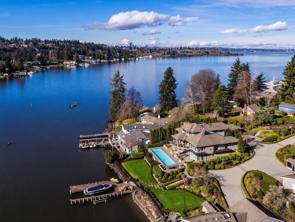This high-angle aerial view captures a luxurious waterfront estate situated on a serene lake, showcasing a multi-level residence with a prominent rectangular swimming pool. The property features expansive green lawns, a private dock with a boat, and a winding driveway, all set against a backdrop of lush trees and a distant city skyline. The perspective emphasizes the home's prime location and seamless integration with the natural landscape.