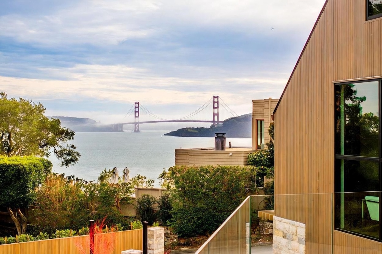 This elevated perspective captures a modern home's balcony, featuring a glass railing that offers an unobstructed view of the Golden Gate Bridge across the bay. The exterior of the house is clad in vertical light wood siding, complemented by large windows that reflect the surrounding landscape. Lush greenery and trees frame the foreground, creating a serene and picturesque setting that emphasizes the property's prime coastal location.