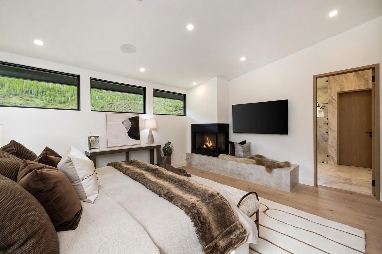 This primary bedroom features a contemporary design with a neutral color palette and a cozy, textured aesthetic. A central fireplace with a stone hearth serves as a focal point, positioned next to a wall-mounted television and a doorway leading to an en-suite bathroom. Three horizontal windows provide natural light and views of the surrounding landscape, while a plush bed with layered linens and a faux-fur throw adds warmth to the space.
