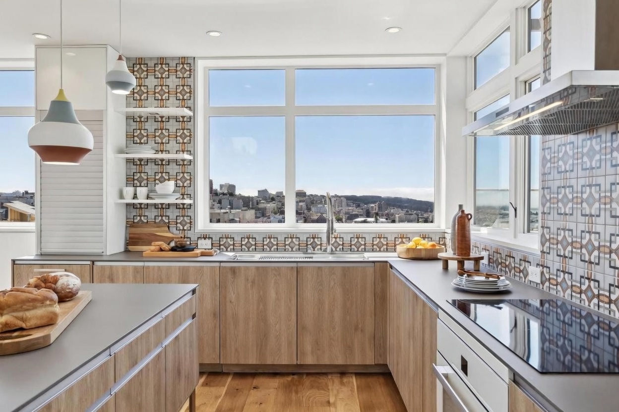 This modern kitchen features light wood cabinetry paired with matte gray countertops and a distinctive patterned tile backsplash that extends up the wall. A large central window frames a panoramic city view, while pendant lights hang above the island, which is topped with a wooden cutting board and fresh bread. The space is bright and airy, blending contemporary design with functional workspace and an inviting atmosphere.