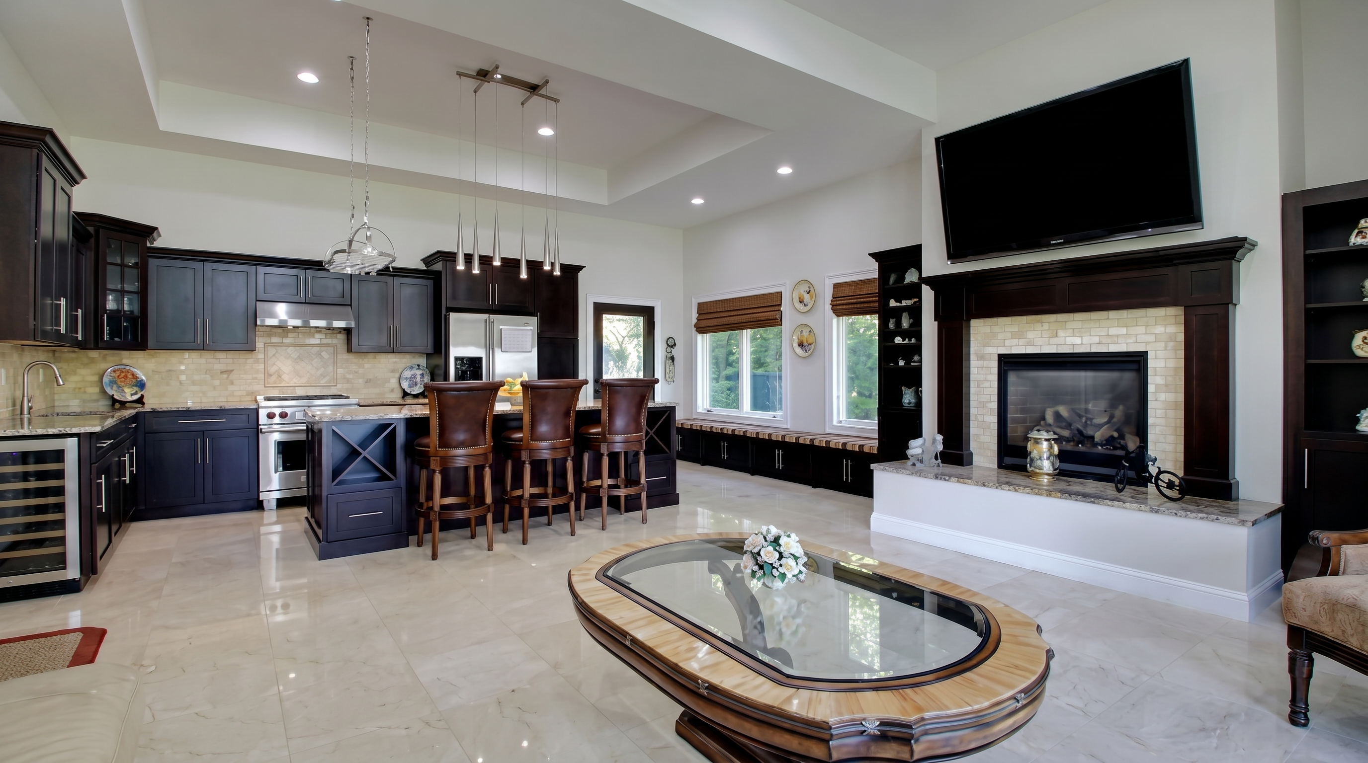 This open-concept space features a modern kitchen seamlessly flowing into a living area. The kitchen is equipped with dark espresso cabinetry, a central island with bar seating, and stainless steel appliances, while the living area showcases a stone fireplace with an integrated television and a unique oval-shaped coffee table. The neutral tile flooring and recessed lighting give the room a sophisticated, high-end, and cohesive appearance.