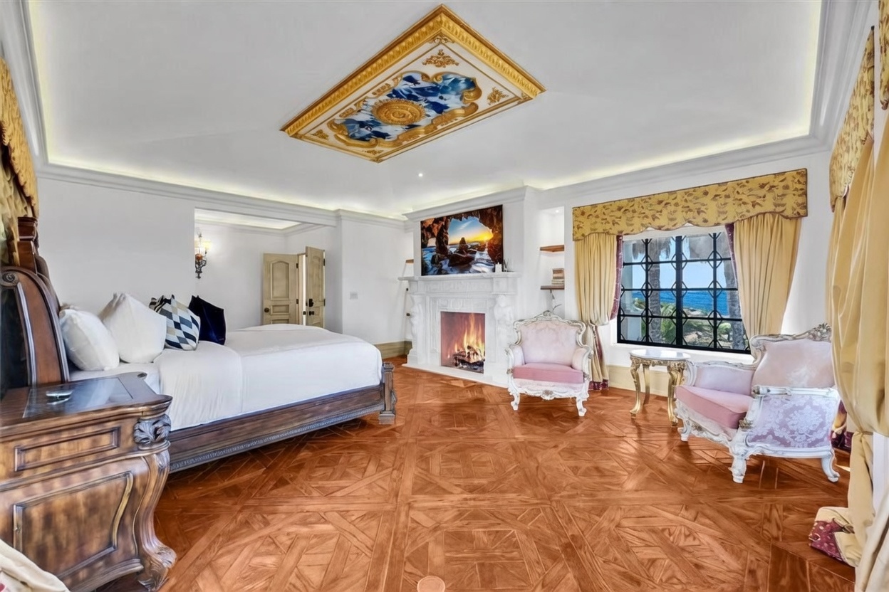 This luxurious primary bedroom features intricate parquet wood flooring and a decorative ceiling medallion with a painted mural. A large bed is positioned near a fireplace with a detailed white mantel, while two ornate pink armchairs sit by a window offering a scenic ocean view. The room is framed by elegant drapery and gold-toned accents, creating a sophisticated and opulent atmosphere.