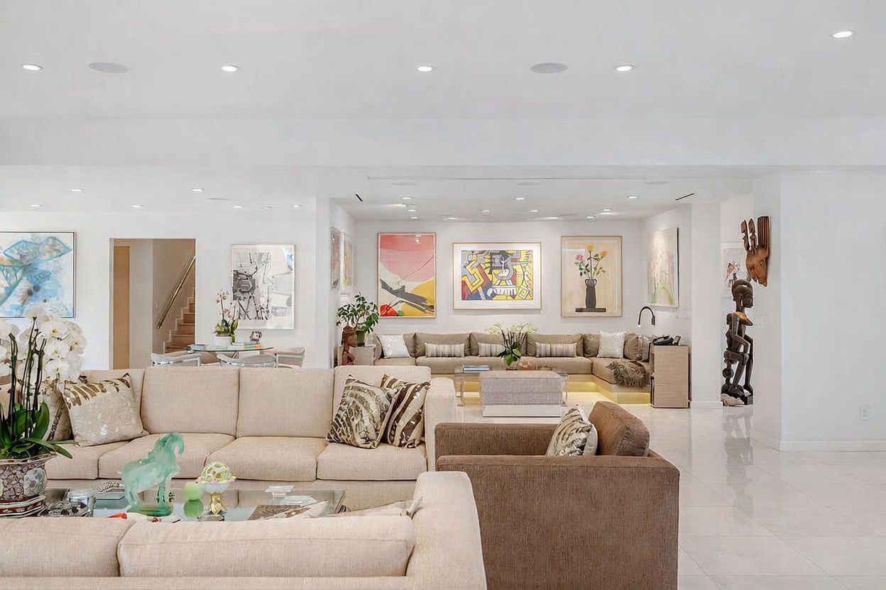 This expansive, open-concept living area features a sophisticated neutral color palette and a bright, airy atmosphere. The space is furnished with multiple large, plush sectionals arranged for comfortable seating, accented by a variety of colorful contemporary artwork on the walls. A tall, dark sculptural piece stands prominently near the right side, while recessed lighting and a polished light-colored floor enhance the modern, gallery-like aesthetic.