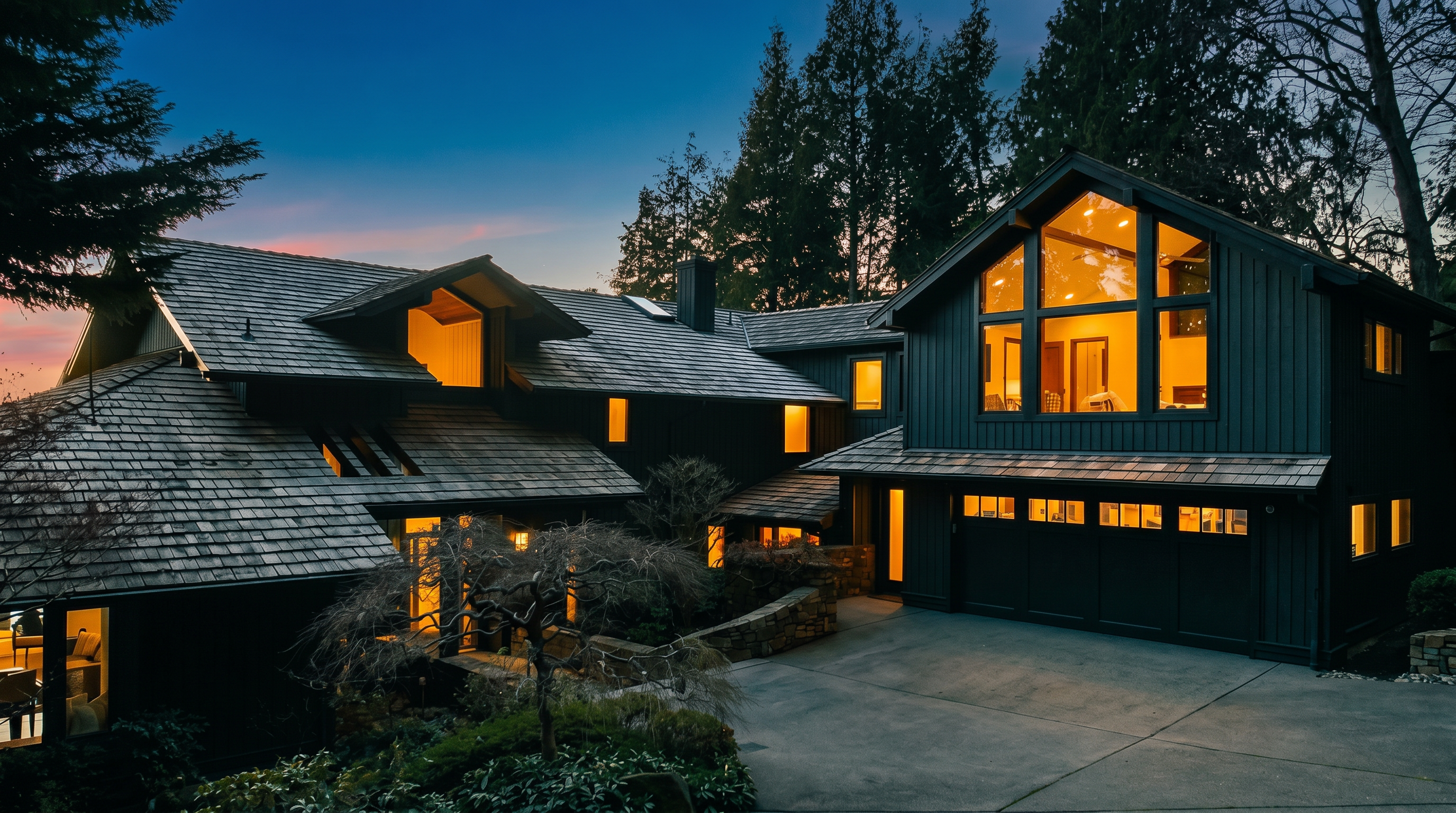 This striking contemporary home features a dark, moody exterior with cedar shake roofing and large, illuminated windows that create a warm, inviting glow against the twilight sky. The property includes a spacious concrete driveway leading to an integrated garage, surrounded by lush, mature landscaping and stone retaining walls. The architectural design emphasizes clean lines and a dramatic, multi-level silhouette that blends modern aesthetics with a natural, wooded setting.