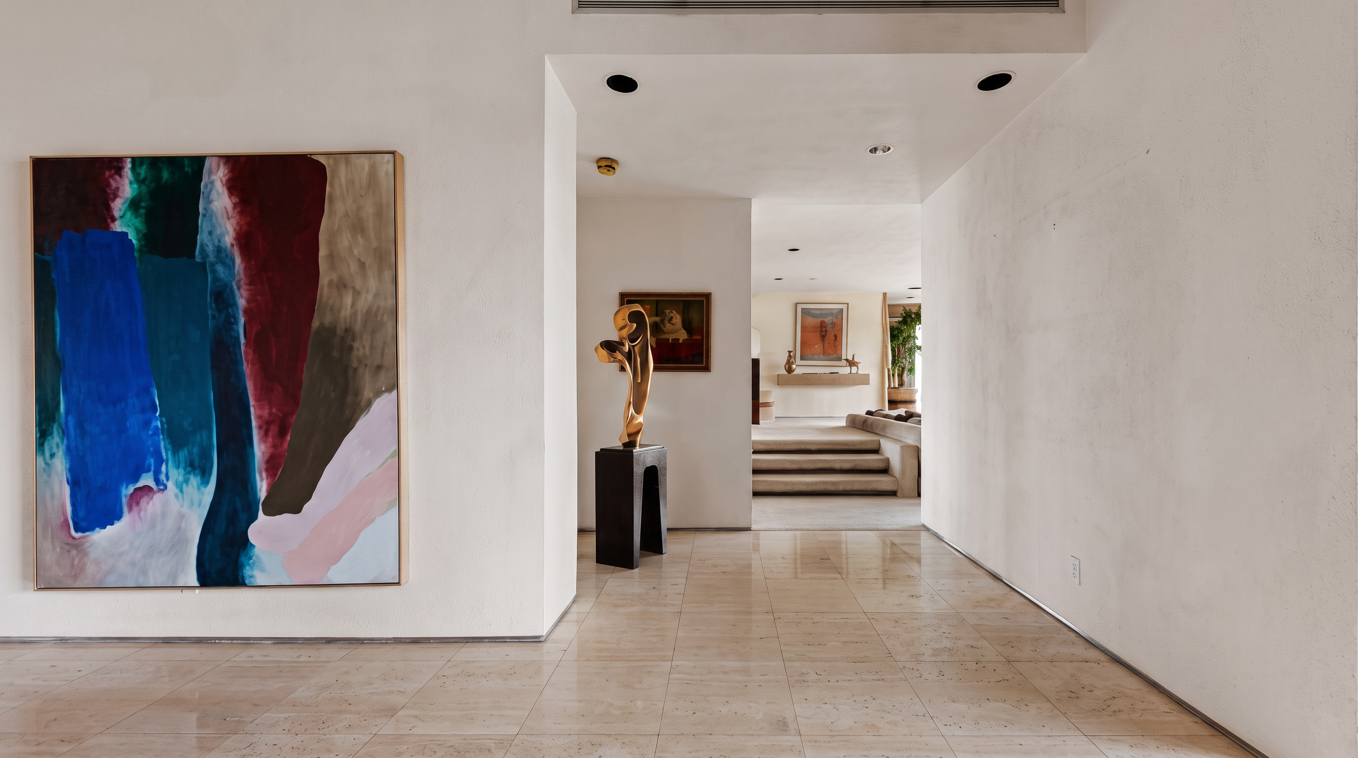 This spacious, minimalist foyer features polished light-toned stone flooring and crisp white walls, creating an airy and sophisticated atmosphere. A large, vibrant abstract painting hangs on the left wall, while a sculptural gold-toned art piece stands on a dark pedestal, leading the eye toward a set of steps that ascend into a bright, open living area. The perspective is centered, emphasizing the clean lines and gallery-like aesthetic of the home's transition space.