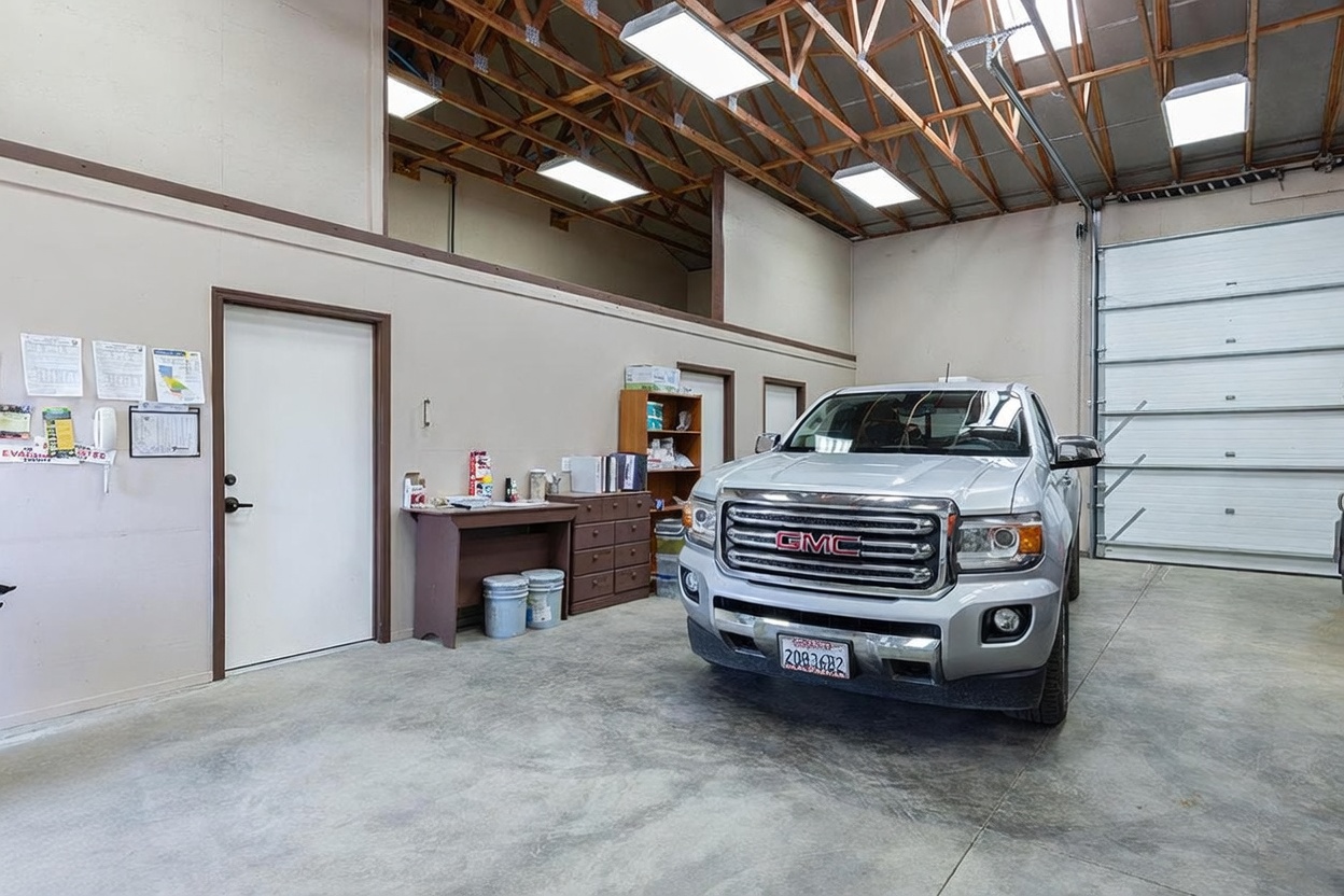 This spacious, high-ceilinged garage features a large roll-up door and a silver GMC pickup truck parked inside. The area includes a functional workspace with a desk, storage shelving, and a white door leading to an adjacent room, all set against a neutral-toned wall. The industrial aesthetic is enhanced by exposed wooden rafters and bright overhead lighting, creating a practical and versatile storage or workshop environment.