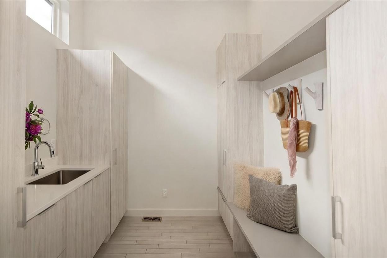This functional laundry and mudroom area features light wood-toned cabinetry and a built-in bench with storage. A stainless steel sink is integrated into the counter on the left, while the right side provides hooks for hanging items and a cushioned seating area. The space is finished with light-colored tile flooring and clean, neutral walls, creating a bright and organized utility room.