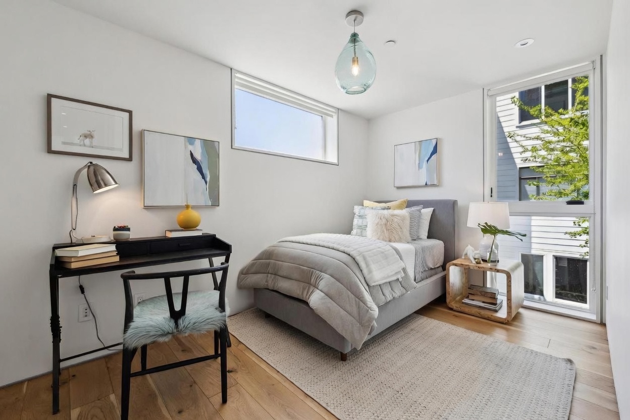 This bright and modern bedroom features a neutral color palette with light wood flooring and a textured area rug. The room is furnished with a single bed, a sleek black desk with a matching chair, and a unique curved side table. Large windows provide ample natural light and a view of the exterior, while a decorative glass pendant light adds a contemporary touch to the space.
