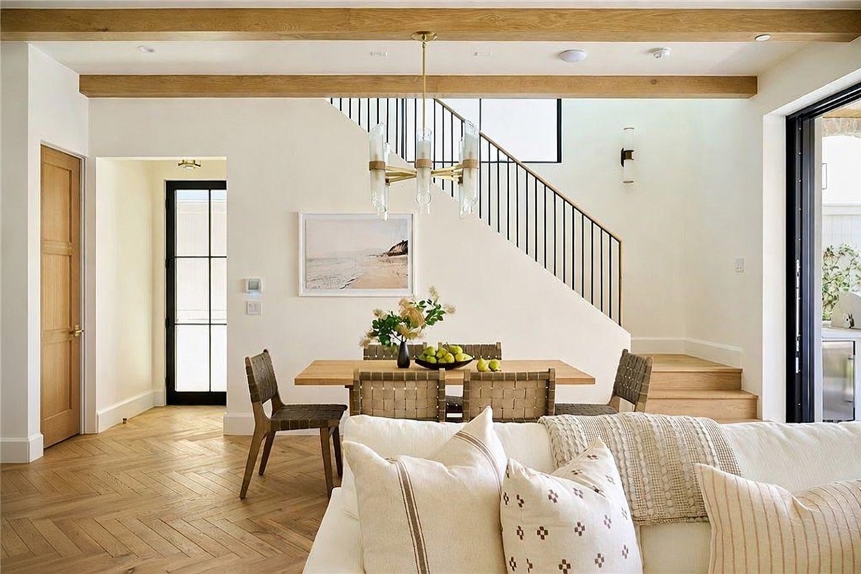 This bright and airy dining area features a light wood table surrounded by woven chairs, positioned beneath a modern multi-light chandelier. The space is anchored by a staircase with a black metal railing and light wood steps, while a large framed landscape print hangs on the wall. The room showcases a cohesive design with light wood flooring in a herringbone pattern and a neutral color palette that creates an inviting, open-concept atmosphere.