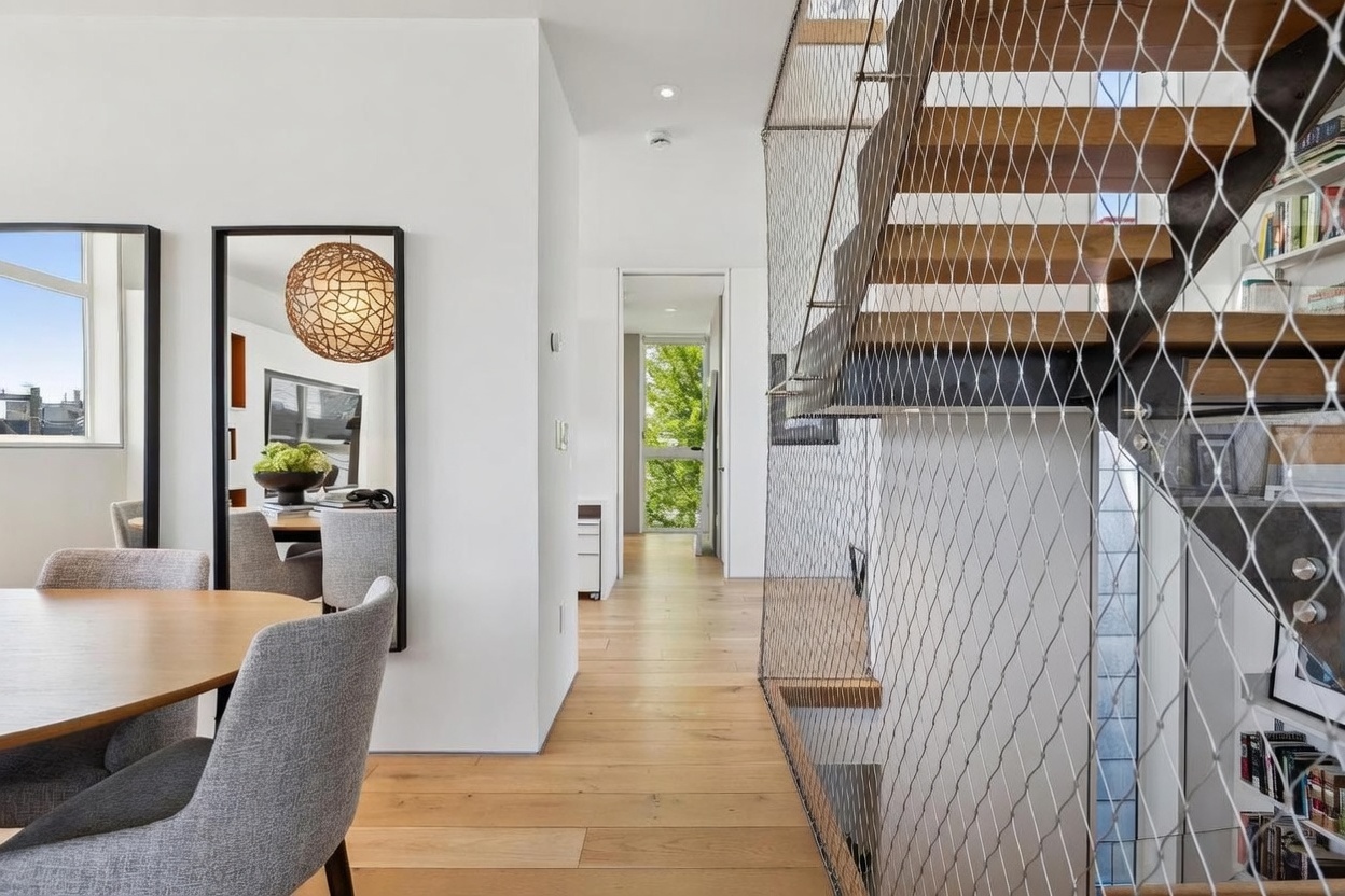 This modern interior space features a light wood floor that transitions from a dining area into a bright hallway leading toward a windowed view of greenery. To the right, a striking staircase is enclosed by a unique metal mesh safety screen, adding an industrial-chic aesthetic to the open-plan design. The perspective captures the clean lines and minimalist architecture, emphasizing the airy, light-filled atmosphere of the home.