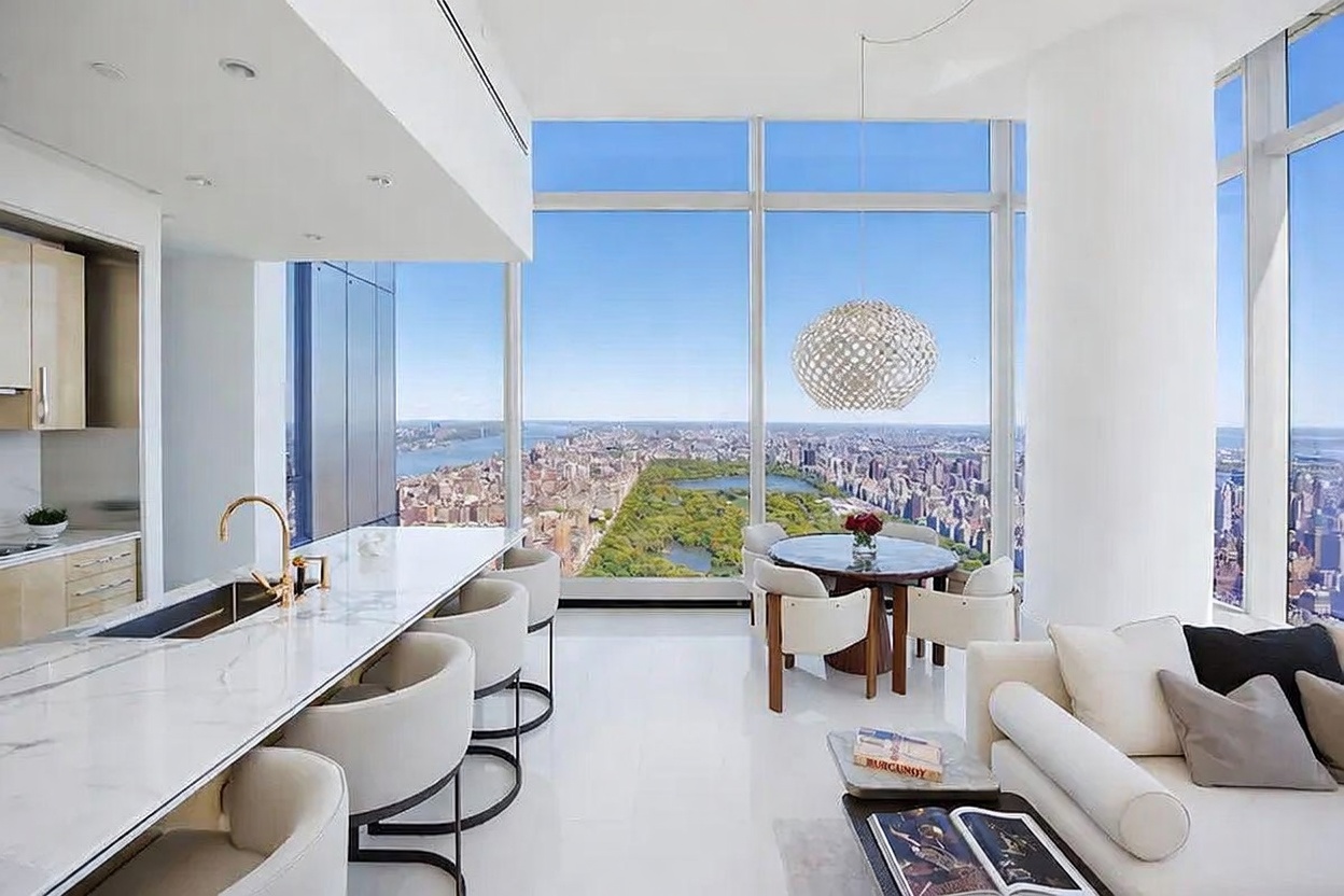 This high-rise interior space features an open-concept kitchen and dining area defined by expansive floor-to-ceiling windows that offer a panoramic view of Central Park and the surrounding city skyline. The kitchen is equipped with a long stone-topped island with integrated seating, while a circular dining table sits nearby under a modern, spherical pendant light. The bright, minimalist aesthetic is enhanced by white flooring and sleek cabinetry, creating a sophisticated and airy atmosphere.