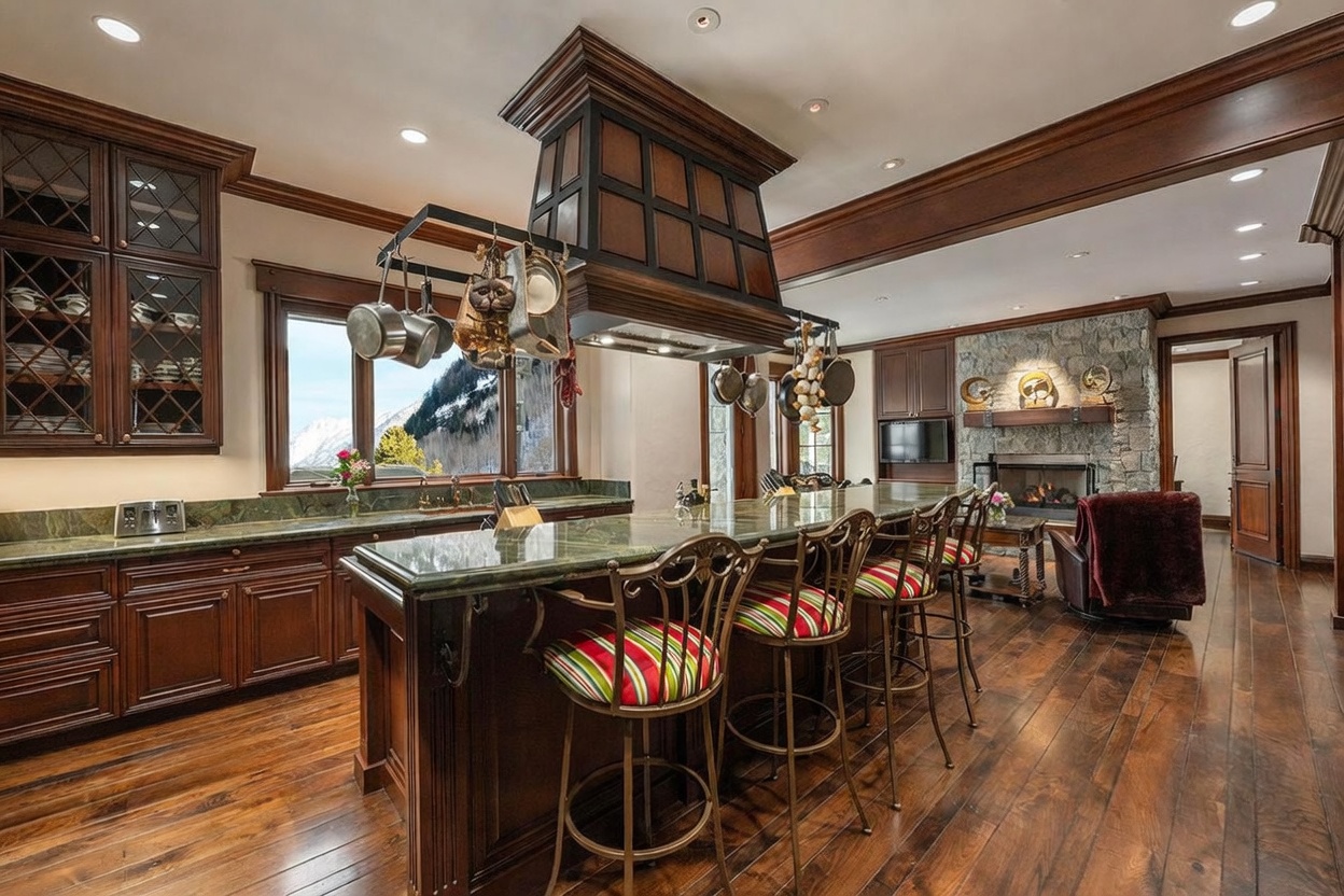 This spacious, rustic-inspired kitchen features dark wood cabinetry and a large island topped with green stone, providing ample seating with striped bar stools. A prominent, custom-built wooden range hood hangs above the island, while a stone-clad chimney breast with a fireplace serves as a focal point in the adjacent living area. The room is bathed in natural light from large windows that frame a scenic mountain view, creating a warm and inviting atmosphere.