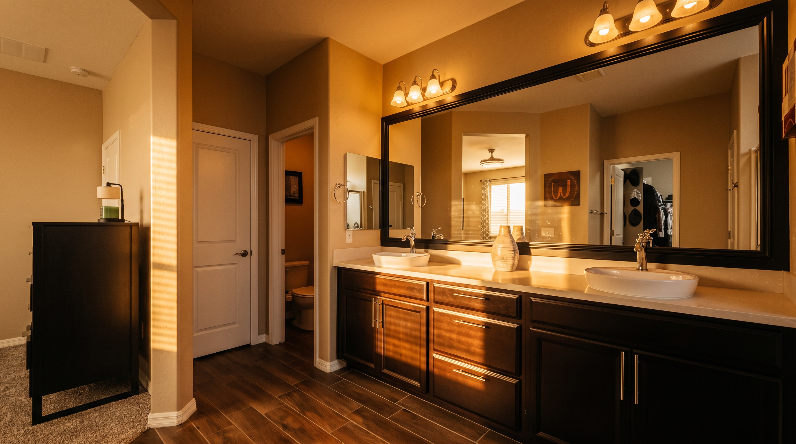 This primary bathroom features a long, dark-toned vanity with dual vessel sinks and a large, framed mirror spanning the length of the counter. The space is finished with wood-look tile flooring and neutral wall paint, creating a clean and contemporary aesthetic. A separate door leads to a private water closet, while the open layout provides a seamless transition from the adjacent carpeted area.