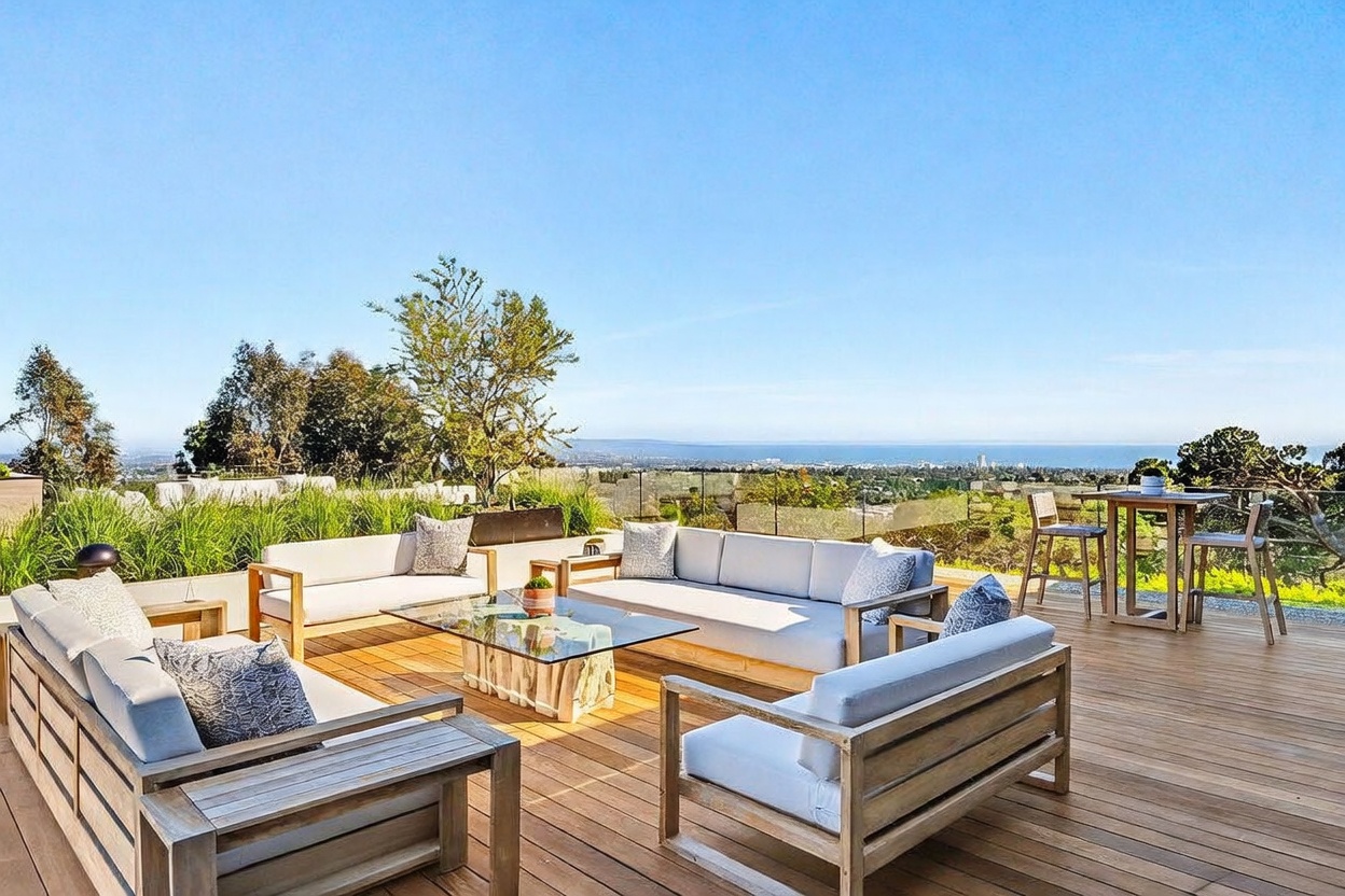 This expansive wooden deck offers a luxurious outdoor living space with a panoramic view of the coastline and horizon. The area is furnished with comfortable light-toned sofas arranged around a glass-topped coffee table, complemented by a small bistro set for dining. A sleek glass railing ensures an unobstructed view, creating a sophisticated and serene atmosphere perfect for entertaining.