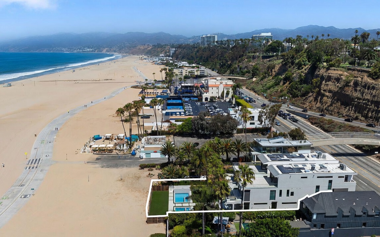 This high-angle aerial view captures a contemporary multi-level residence situated directly along the coastline, highlighted by a white outline. The property features a private swimming pool, a manicured lawn, and expansive flat roof terraces that offer unobstructed views of the Pacific Ocean and the adjacent sandy beach. The surrounding area includes a coastal highway, lush cliffside vegetation, and a nearby beach club, emphasizing the home's prime beachfront location.
