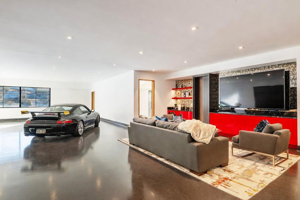 This unique, expansive living space features a polished concrete floor and a high-end sports car parked directly inside the room. The area includes a comfortable seating arrangement with a grey sofa and armchair facing a large wall-mounted television set above a vibrant red media console and wet bar. The room is illuminated by recessed lighting and large windows that provide a view of the outdoors, creating a sleek, modern, and automotive-inspired aesthetic.