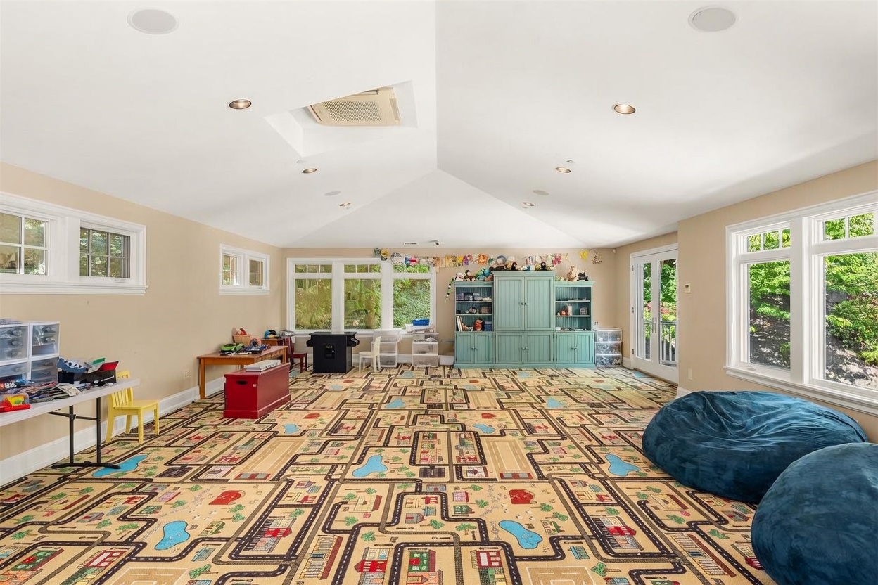 This spacious, light-filled playroom features a unique road-themed carpet, perfect for children's activities. The room is equipped with ample storage, including a large teal cabinet and various shelving units, and is illuminated by recessed lighting and a central skylight. Large windows and glass doors provide plenty of natural light and views of the surrounding greenery, creating an inviting and functional space.