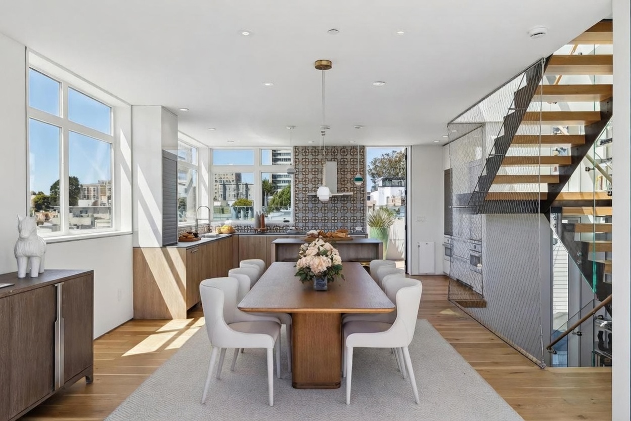 This open-concept dining area features a central wooden table surrounded by six modern white chairs, set atop a neutral area rug. The space flows seamlessly into a kitchen with light wood cabinetry and a patterned tile backsplash, while a staircase with a unique metal mesh railing adds a contemporary architectural element to the right. Large windows provide ample natural light and views of the surrounding urban landscape, creating an airy and sophisticated atmosphere.