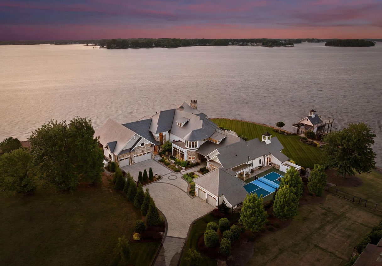 This stunning aerial view captures a sprawling luxury estate situated on a point of land extending into a large lake. The property features a complex roofline, multiple garage bays, a private swimming pool, and a detached dock structure, all set against a vibrant sunset sky. The expansive lawn and paved driveway highlight the grand scale and prime waterfront location of this residence.