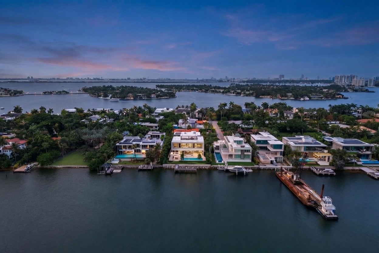 This high-angle aerial view captures a row of modern, multi-story waterfront estates along a calm bay at dusk. The homes feature contemporary architectural lines, expansive glass facades, and private swimming pools overlooking the water, with a large construction barge moored nearby. The scene is set against a backdrop of a distant city skyline under a soft, twilight sky, emphasizing the luxury and exclusivity of the waterfront location.